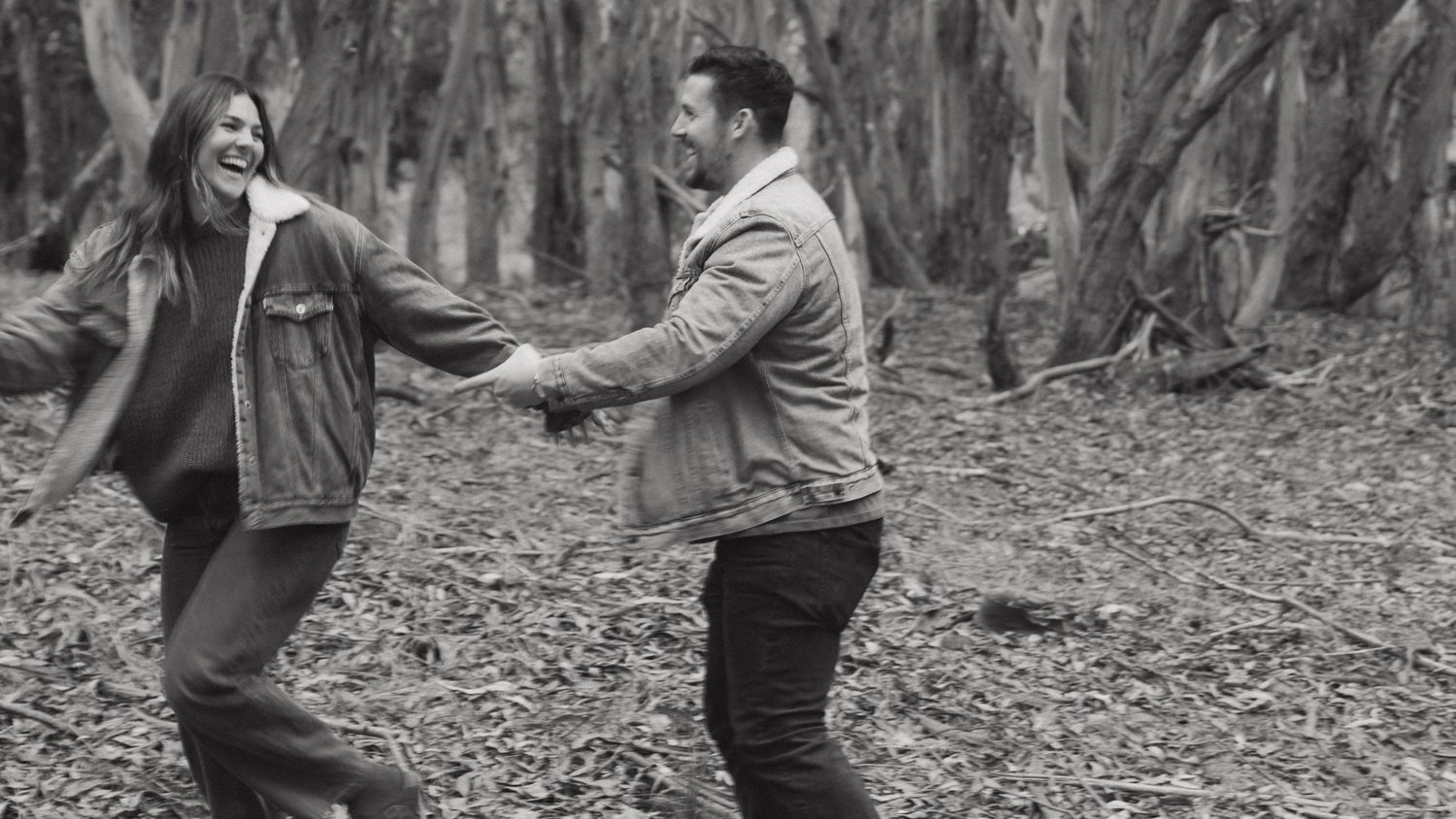 Joyful couple dancing and laughing together in a forest, holding hands in a candid black and white photo.