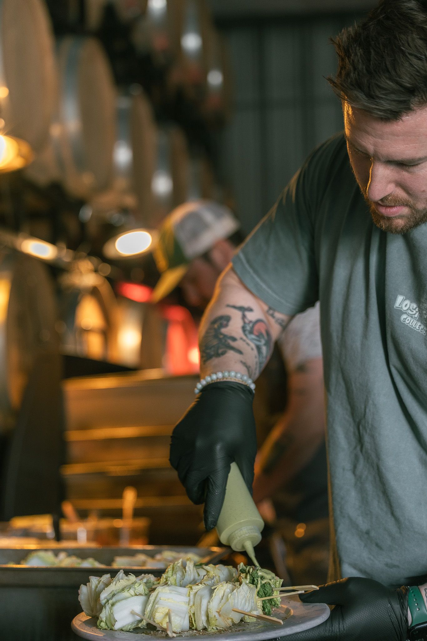 Tattooed chef in black gloves plating food with piping bag at dimly lit restaurant kitchen.
