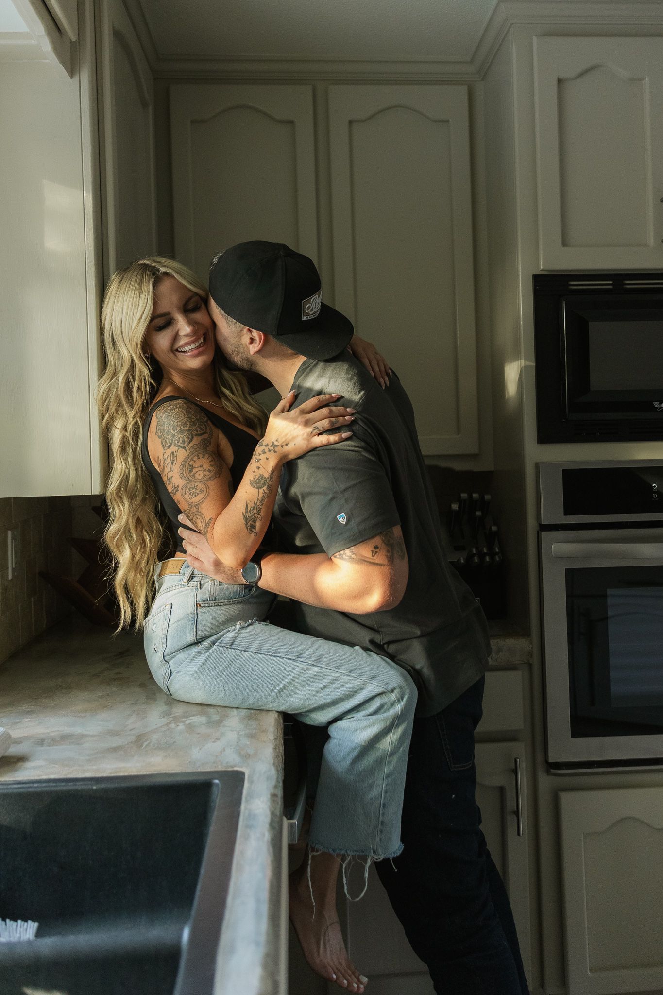Tattooed couple embracing and kissing in a kitchen, woman seated on counter smiling.