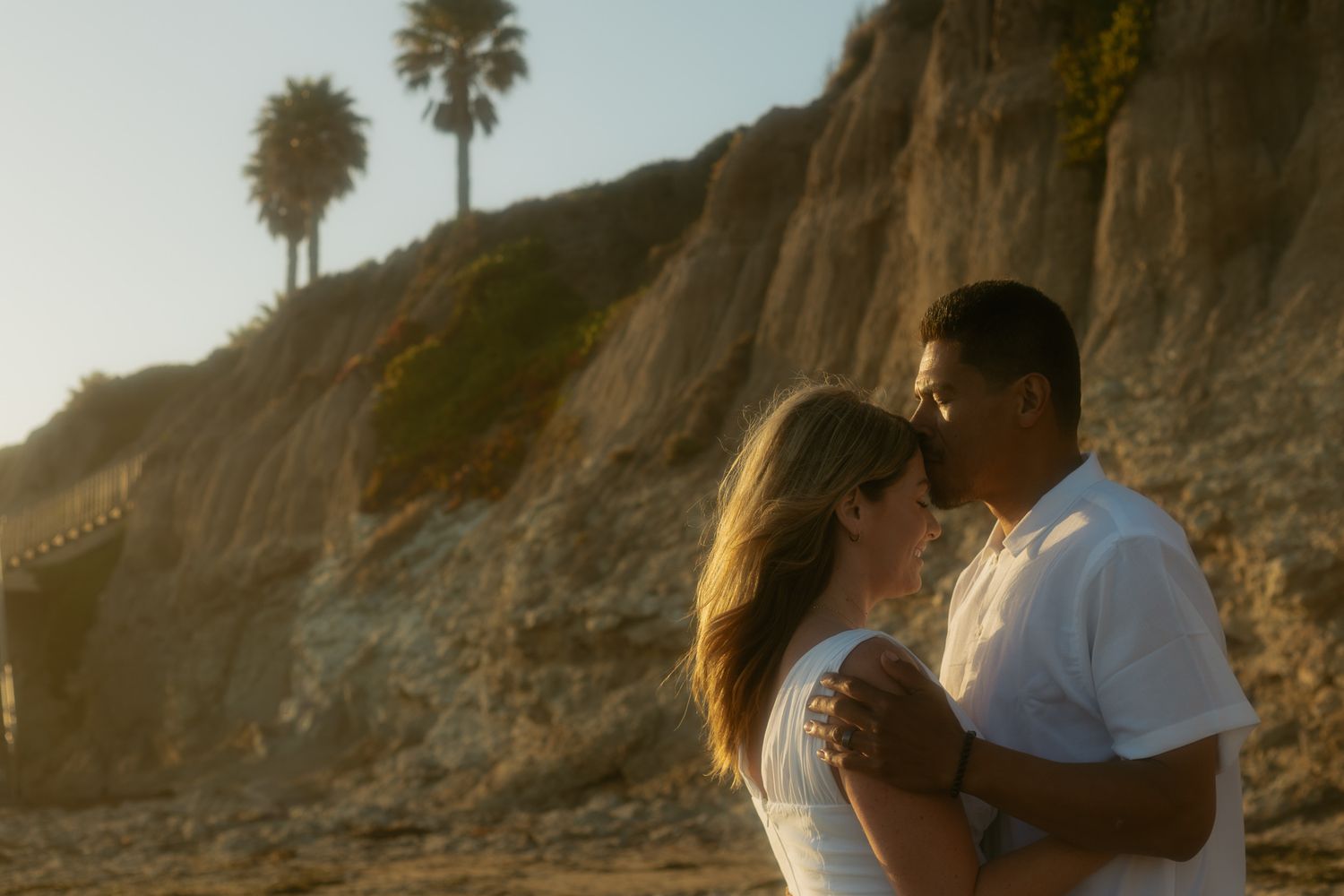 Couple sharing a romantic kiss on a sunny beach with palm trees and sandy cliffs at golden hour in Pismo Beach.