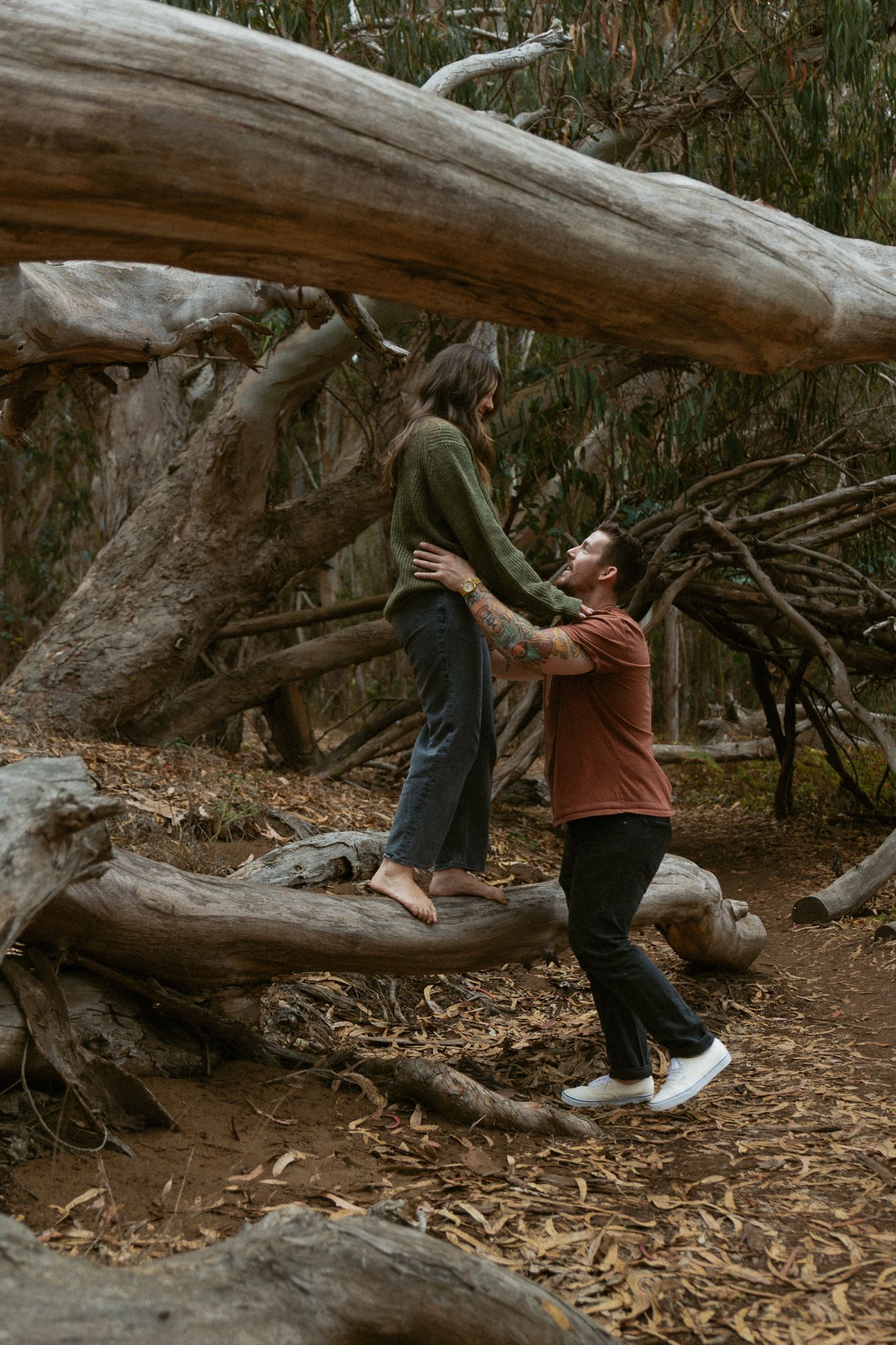 A couple playfully interacts among fallen trees in a forest, woman standing on log while man looks up at her at Montana De Oro State Beach.
