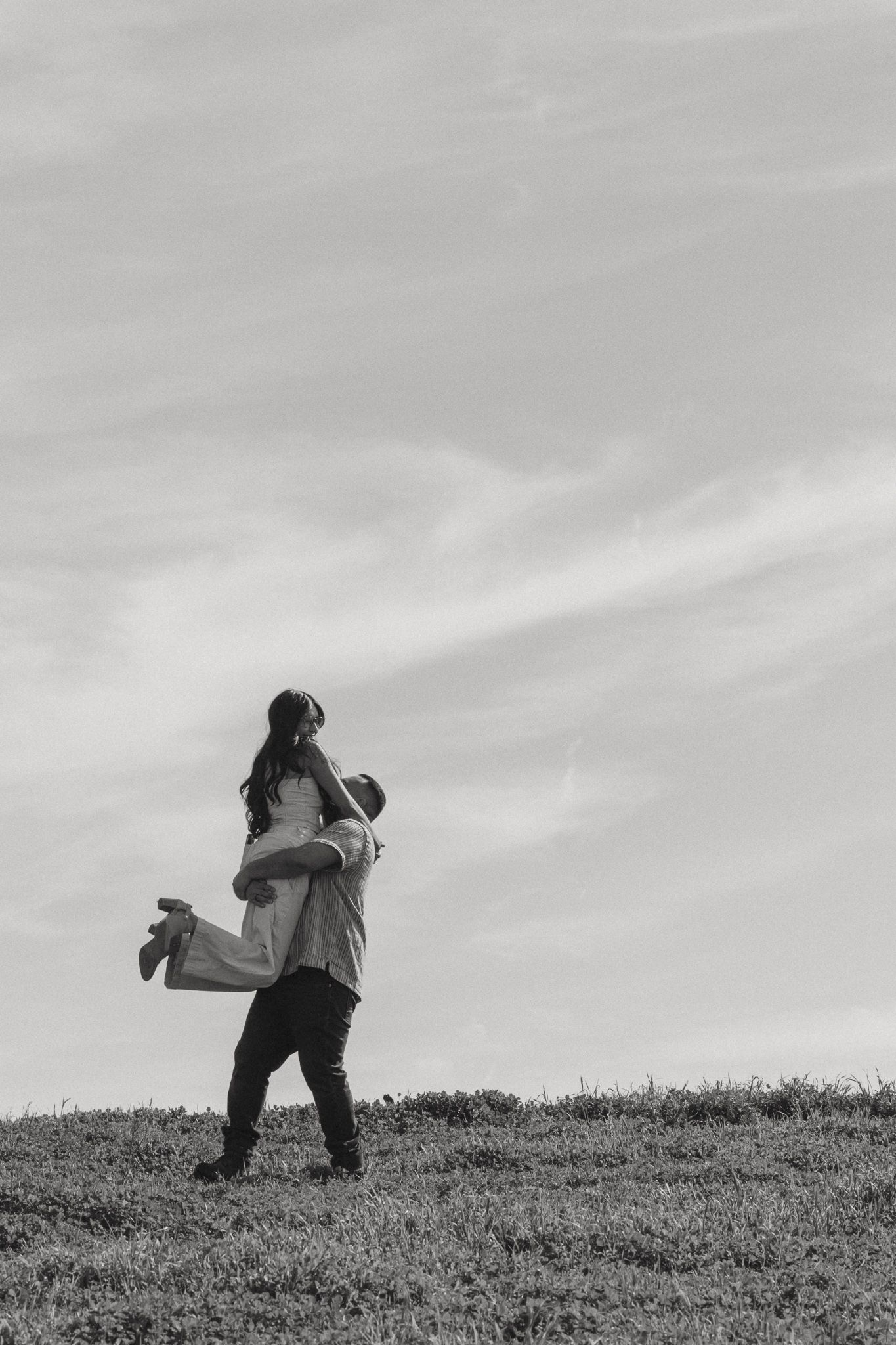 Black and white photo of a man lifting a woman in his arms on a grassy hill against a cloudy sky.