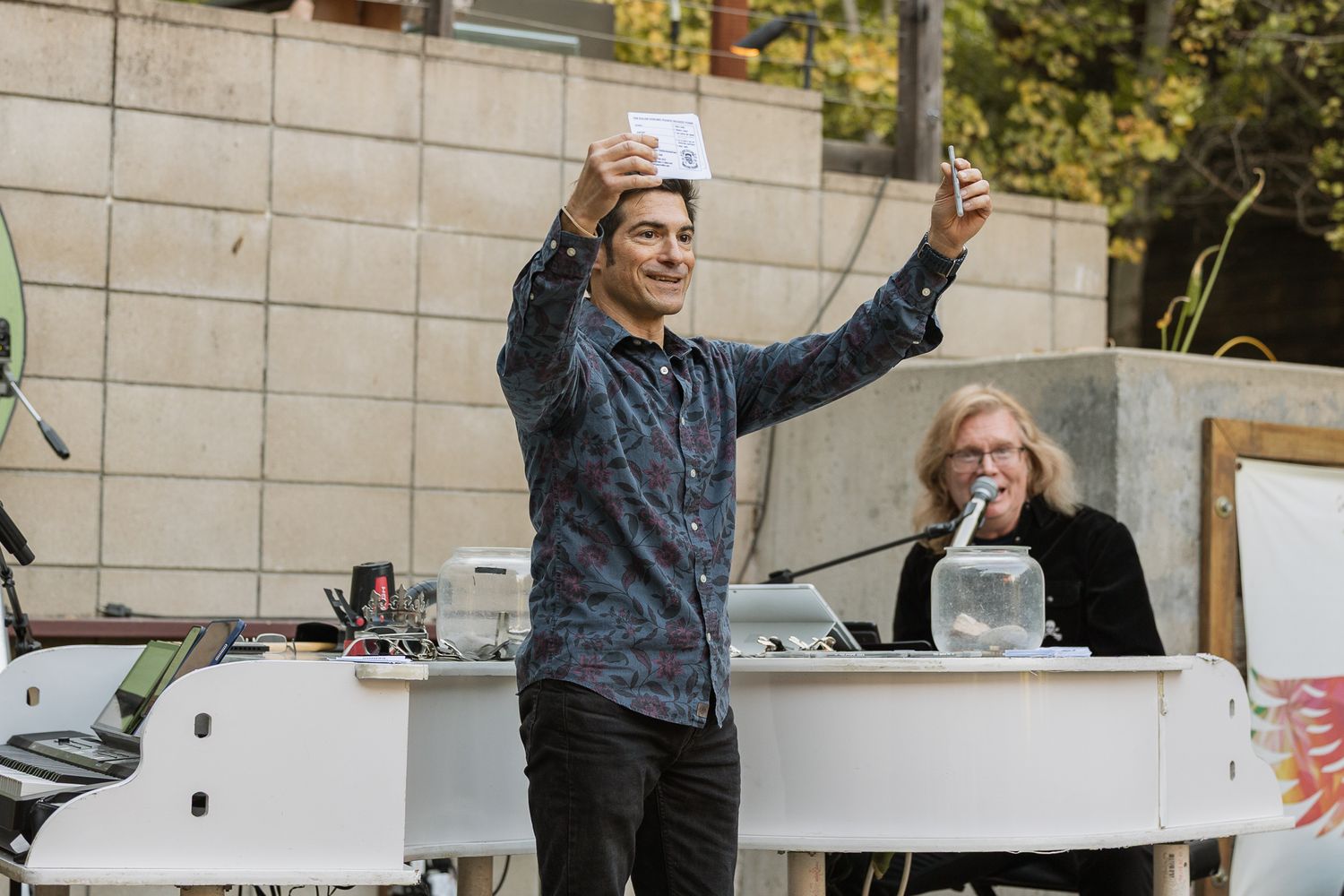 Man taking selfie outdoors at event with white piano and seated woman in background.