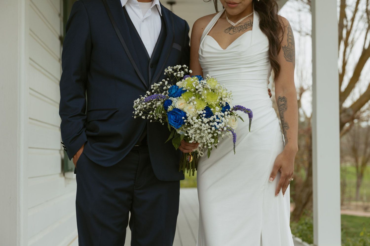 Wedding couple posing on porch, bride in white gown holding blue and white bouquet with groom.