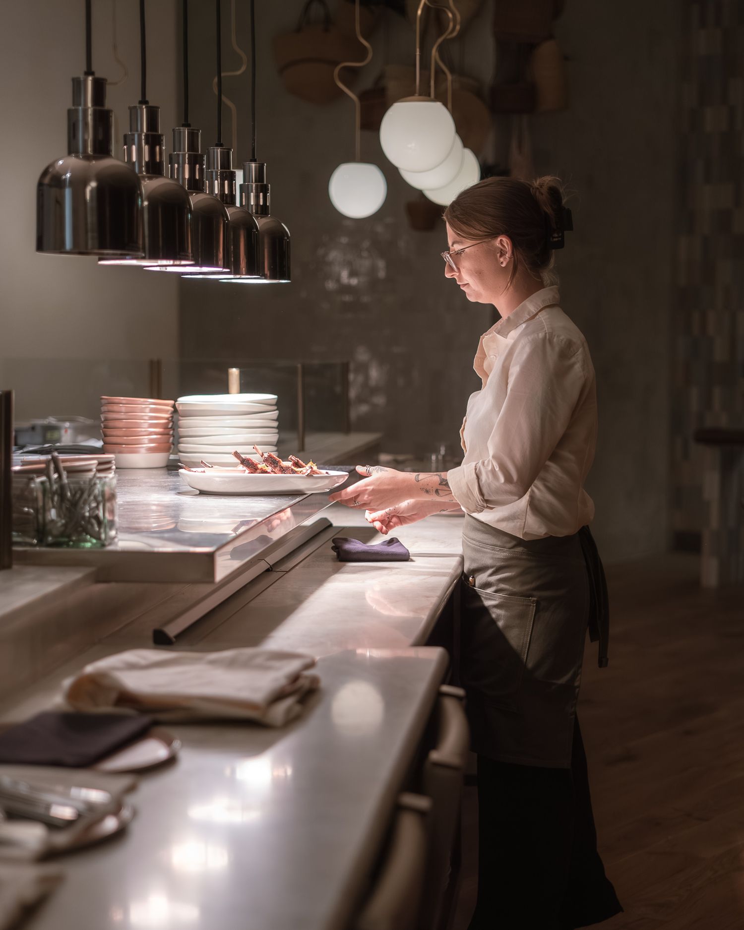 Female chef in apron working at a dimly lit restaurant counter with hanging pendant lights.