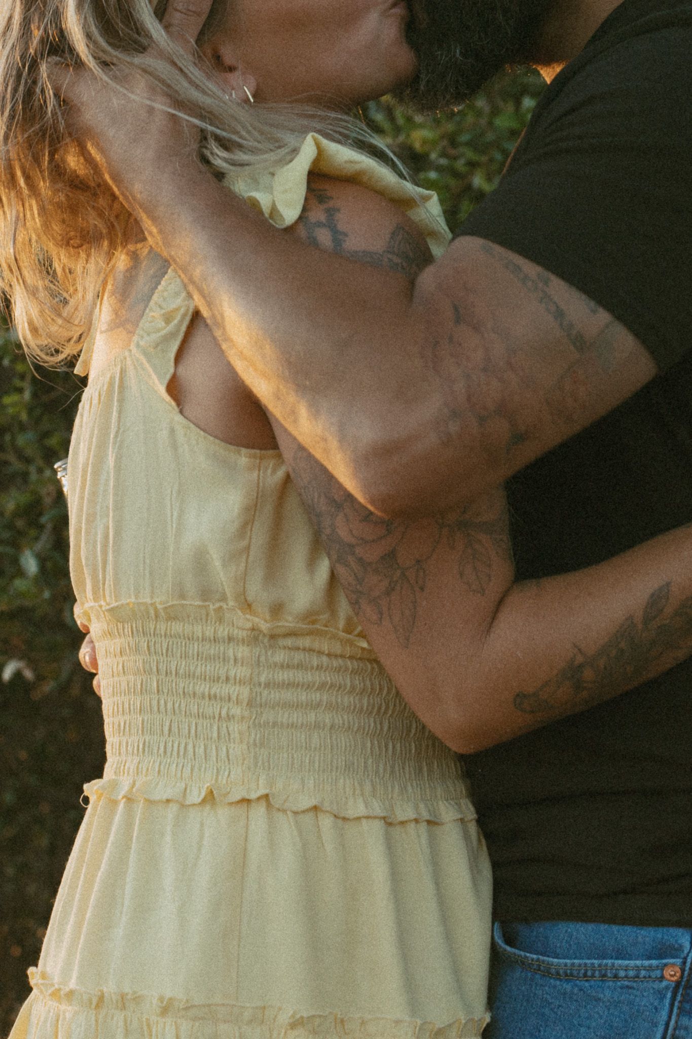 A couple embraces outdoors, the man's tattooed arms wrapped around a woman in a yellow sundress.