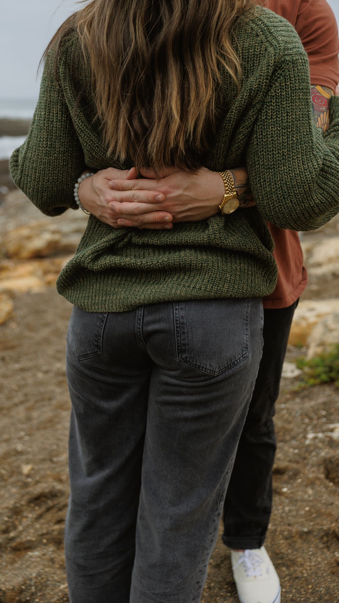 A couple embraces from behind on a rocky shore on Central Coast, hands clasped over a green knit sweater.