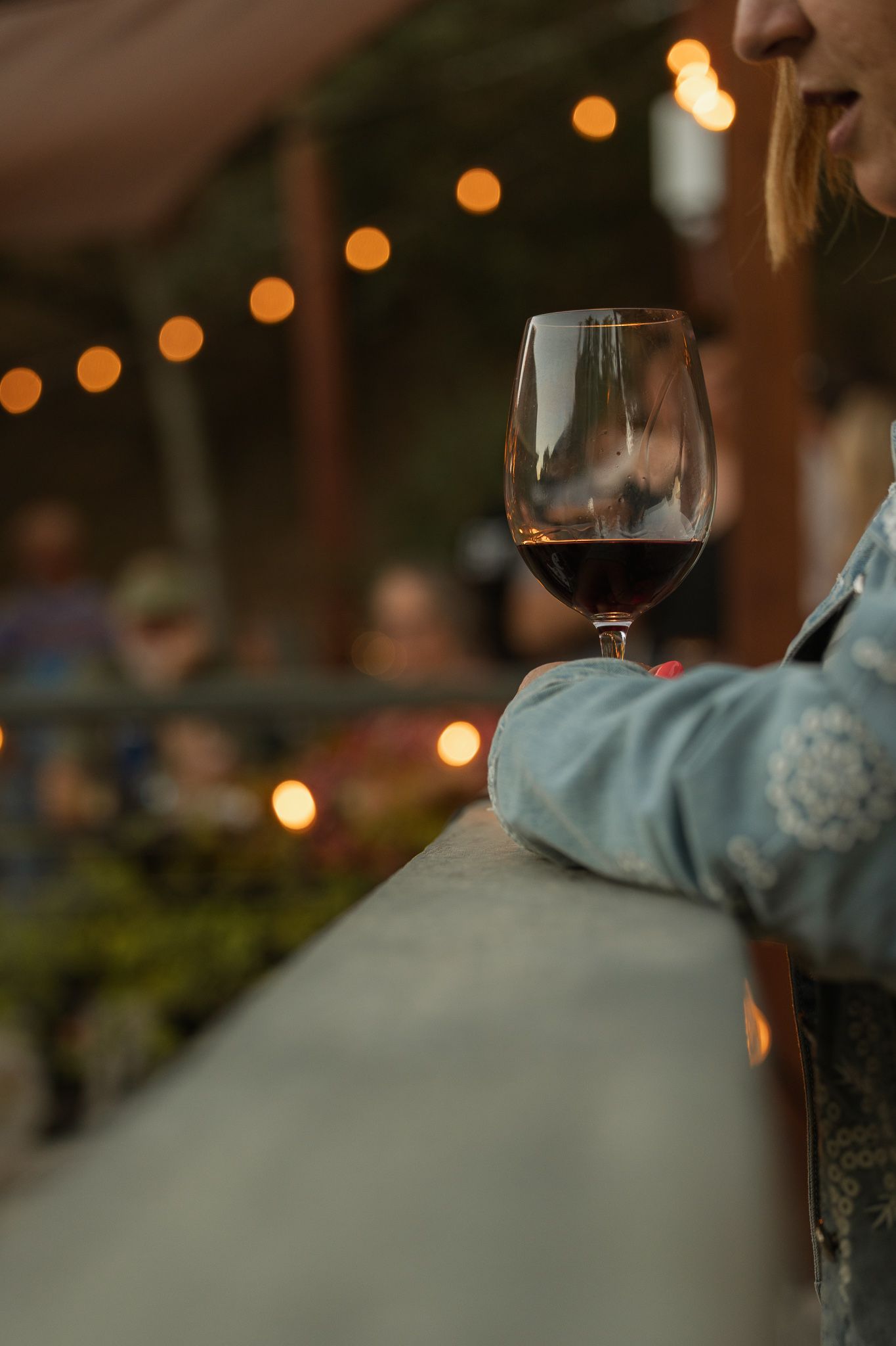 Woman holding a glass of red wine on a balcony with warm string lights glowing in the background at dusk.