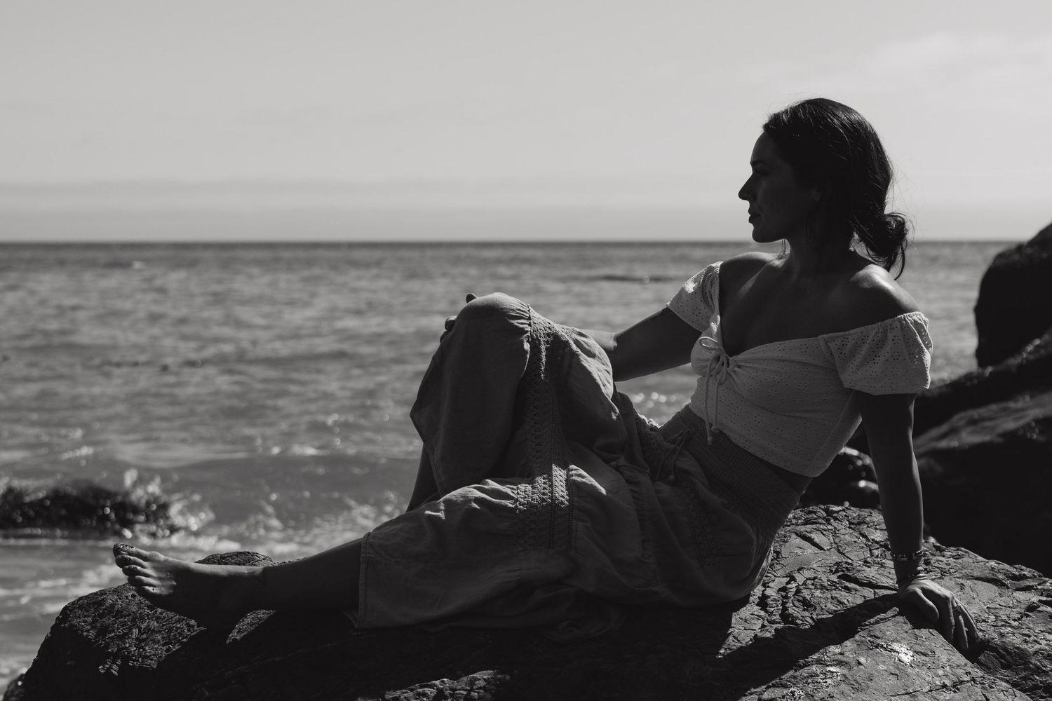 Black and white photo of woman reclining on coastal rocks with ocean waves in background.