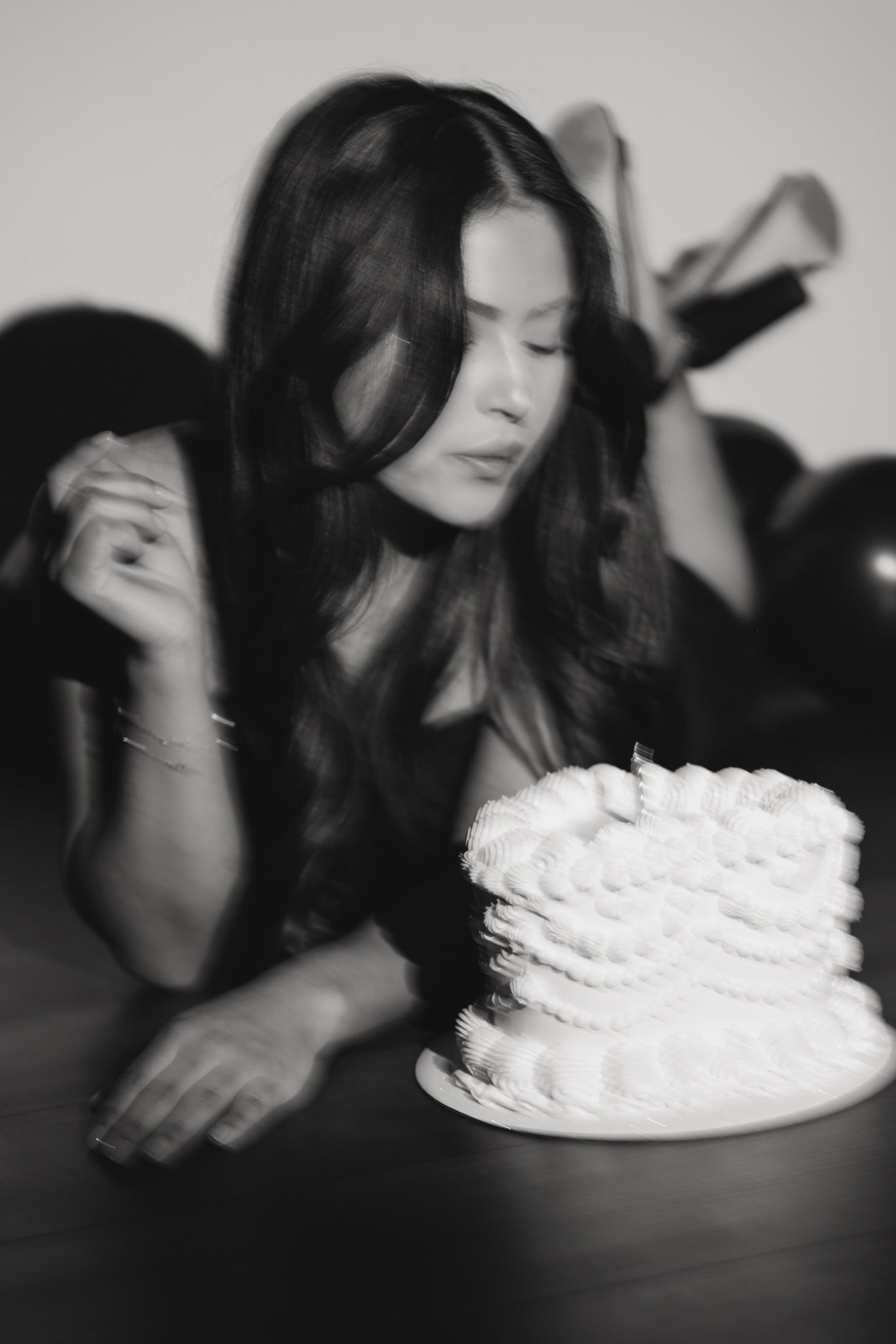 A young woman blows out a candle on a white ruffled cake in a black and white birthday photo.