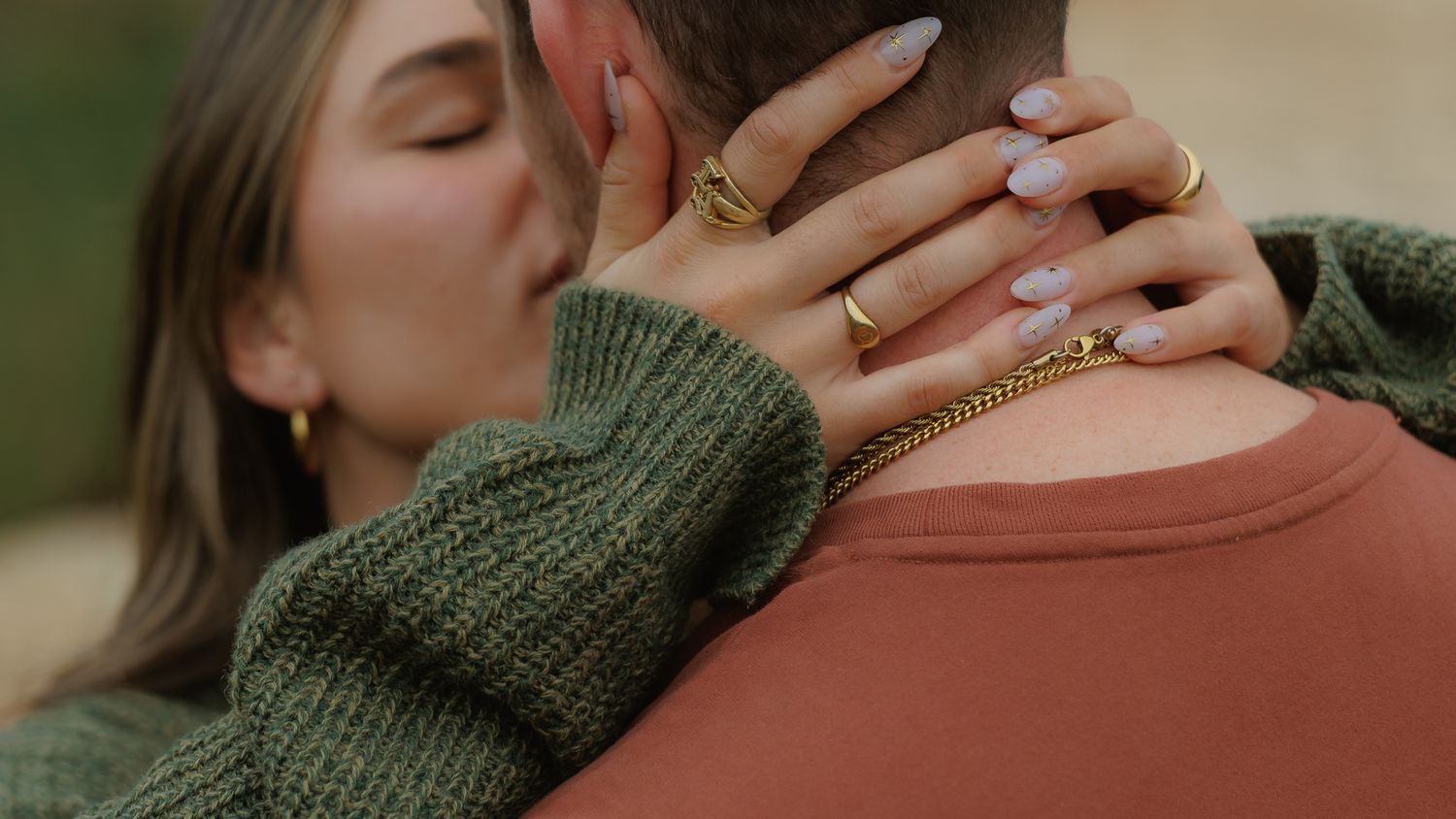 Woman in green sweater kissing man, her hand with gold rings resting on his neck.