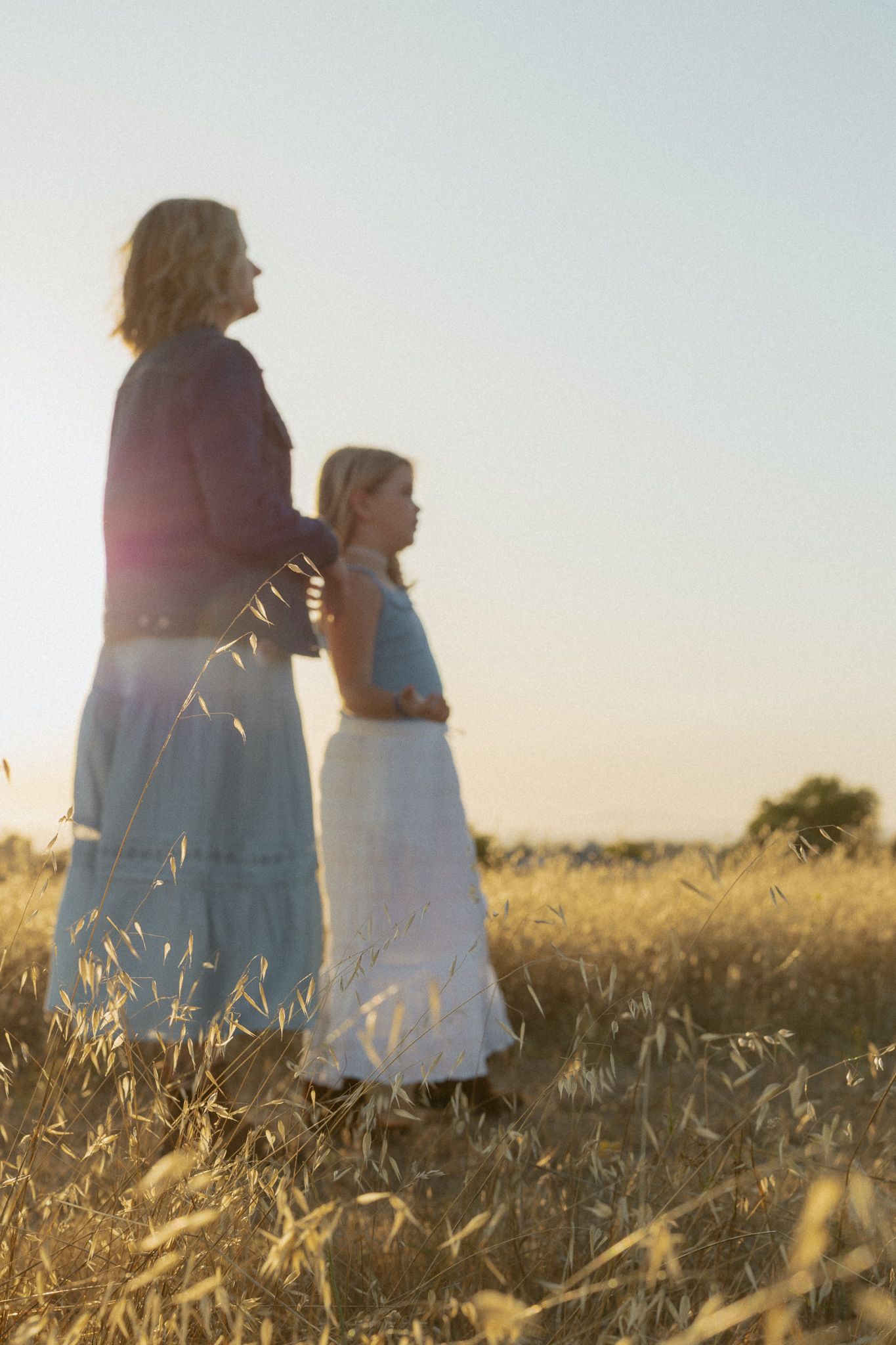 Mother and daughter standing in golden wheat field at sunset wearing flowing skirts.