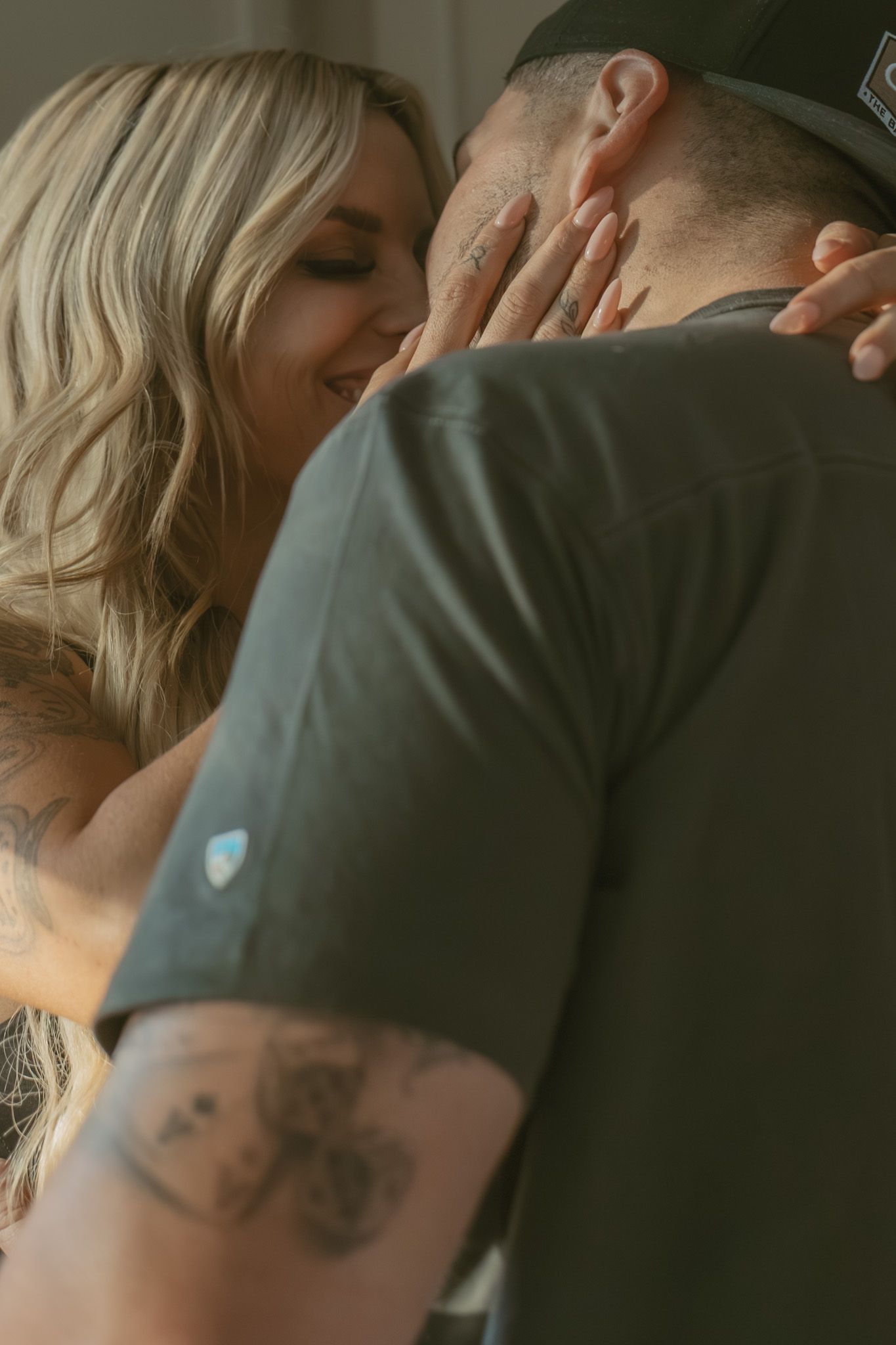 A smiling blonde woman with tattoos tenderly embraces a man in a dark green shirt indoors.