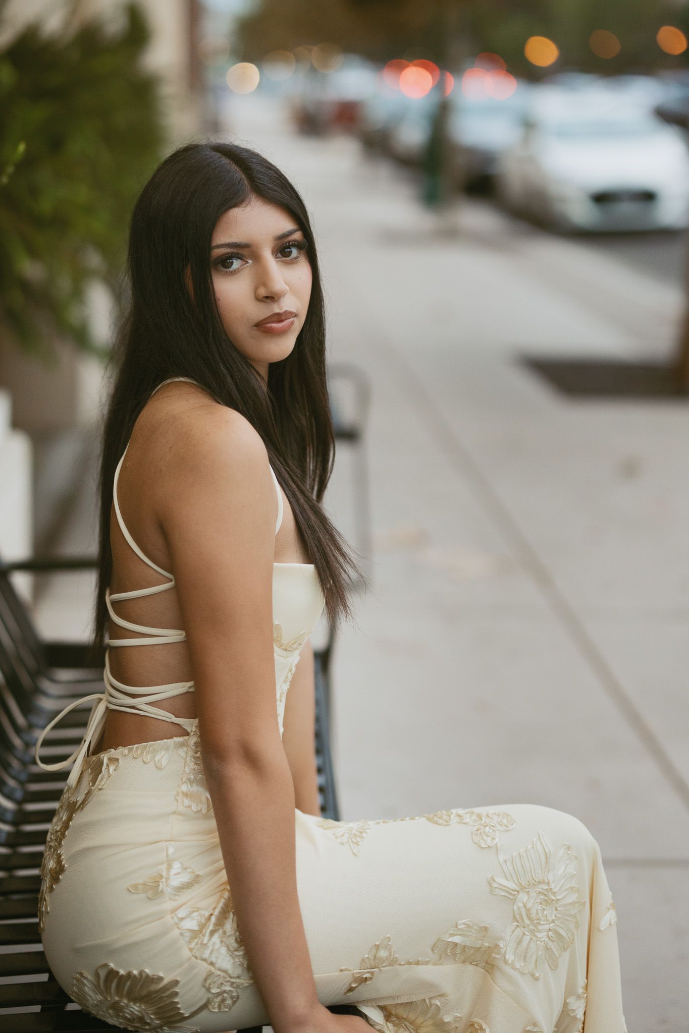 A young woman in a backless floral cream dress sits on a bench along a city sidewalk.