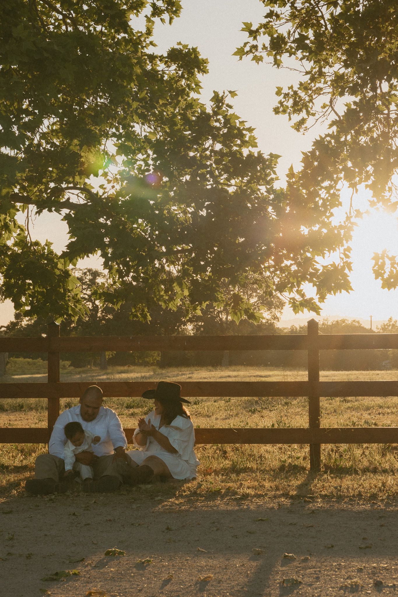 Family of three sitting together by wooden fence under trees in golden sunset light on farm.