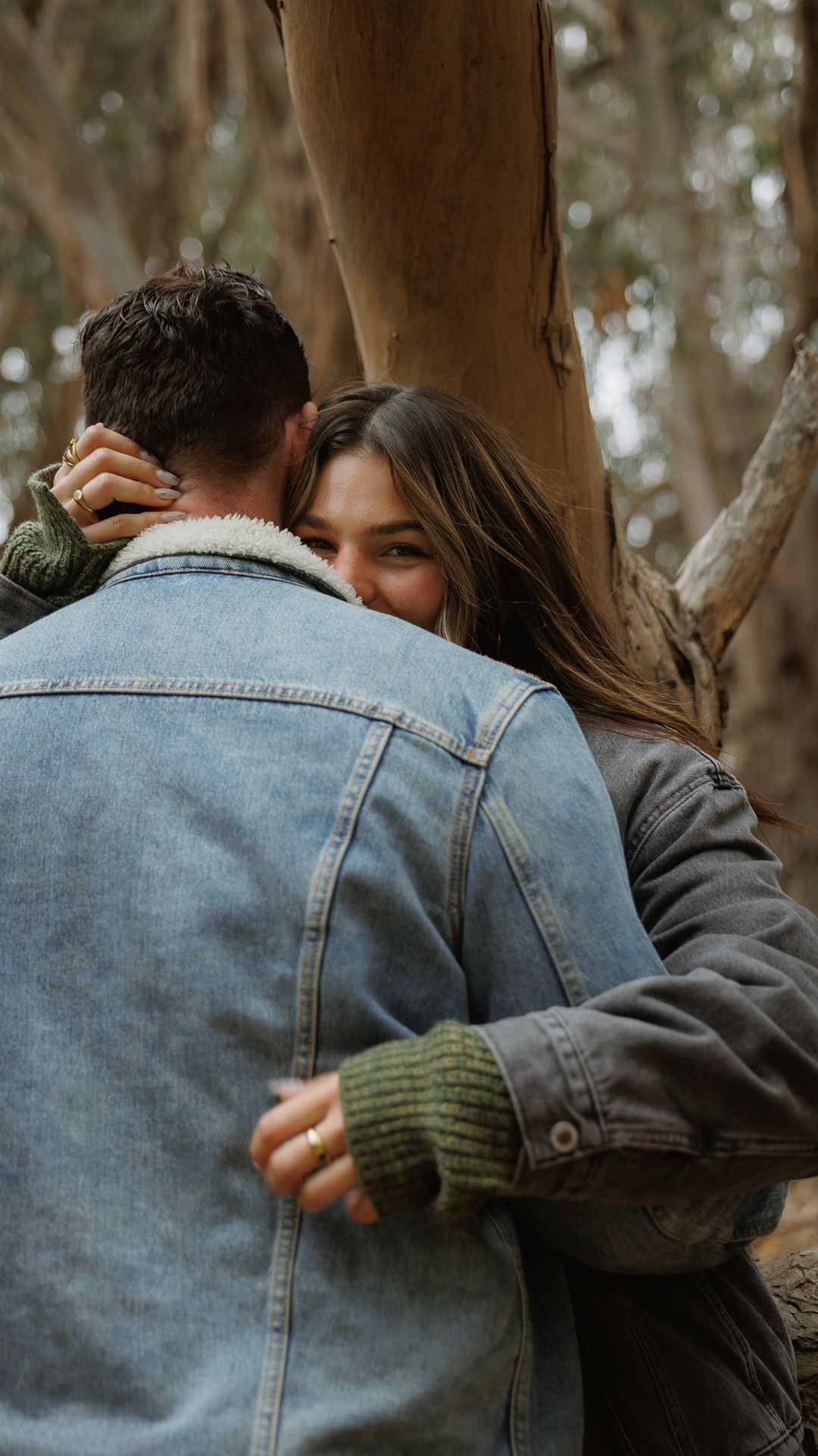 A woman smiles over a mans shoulder as they embrace near eucalyptus trees outdoors.