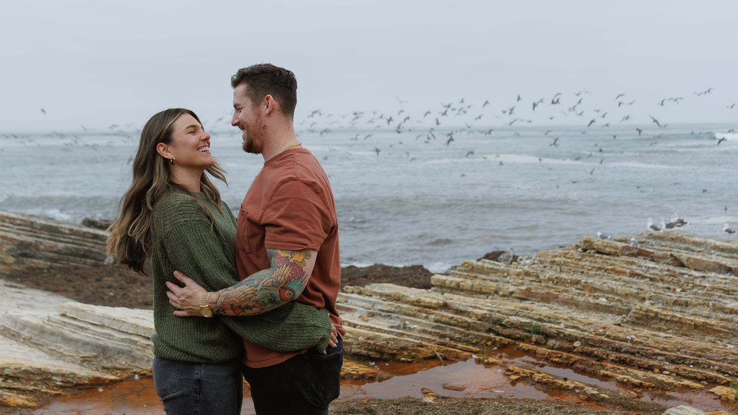 Smiling couple embracing on rocky coastal shore with birds flying over misty ocean.