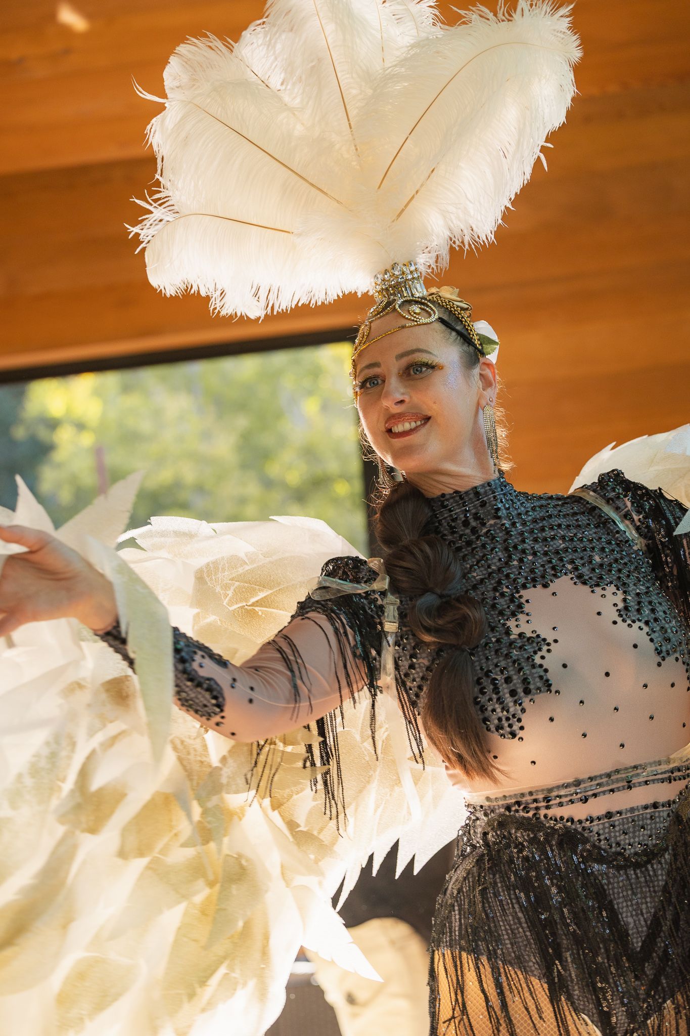 A smiling performer in a black beaded costume with white feather headdress and fans at an outdoor event.