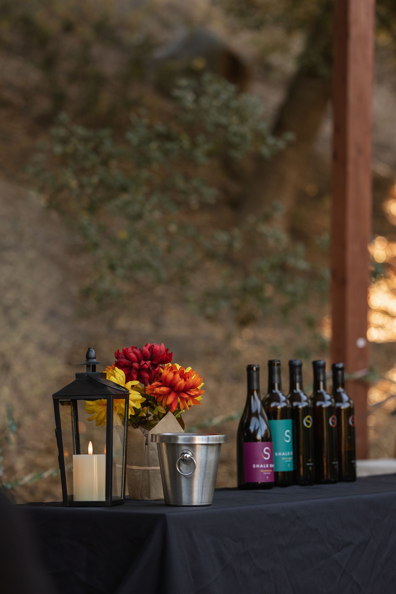 Elegant wine tasting table with candle lantern, fall flowers, ice bucket, and multiple wine bottles on black tablecloth.