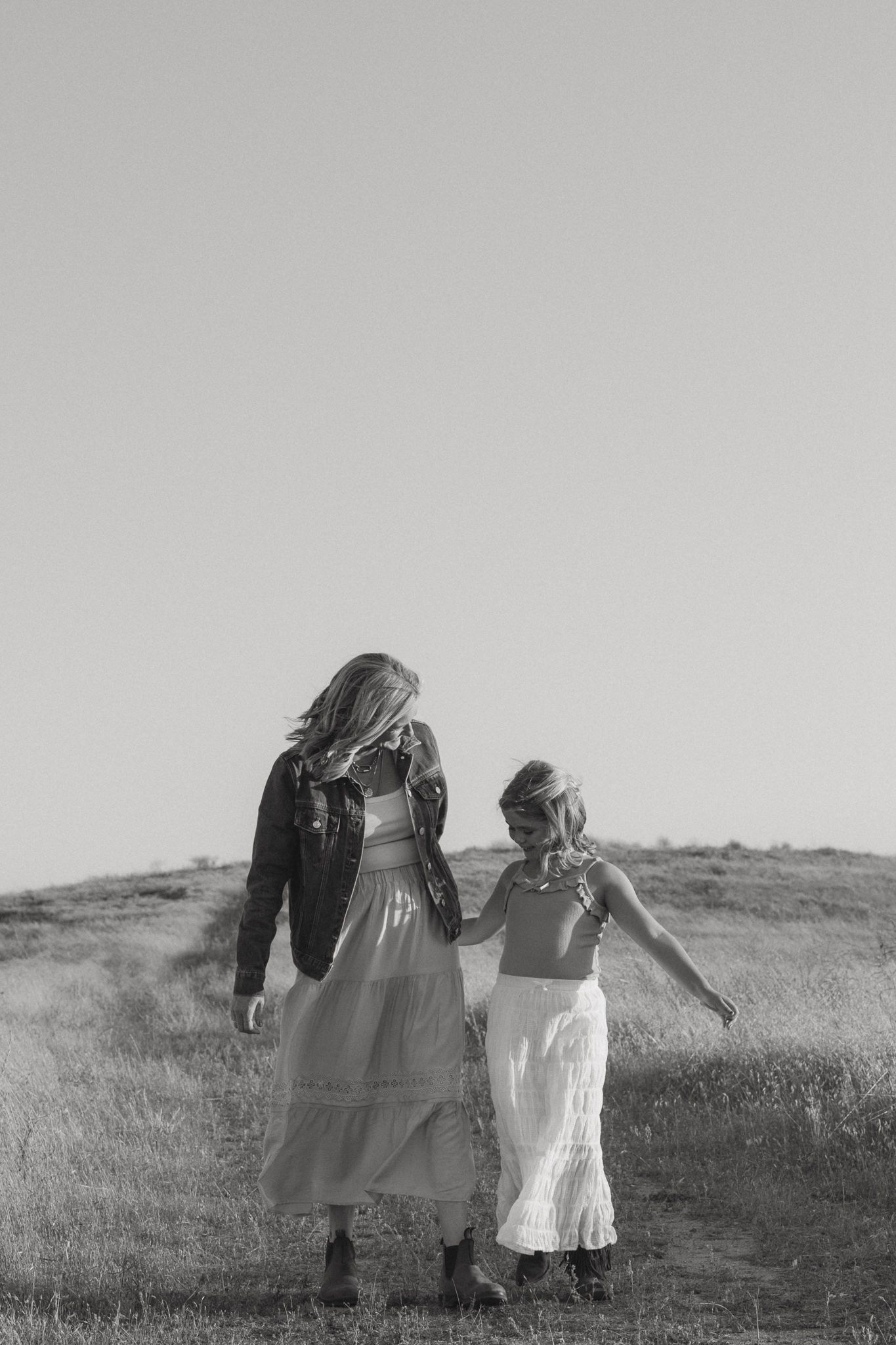 A mother and daughter walk hand in hand through an open field in a black and white photo.