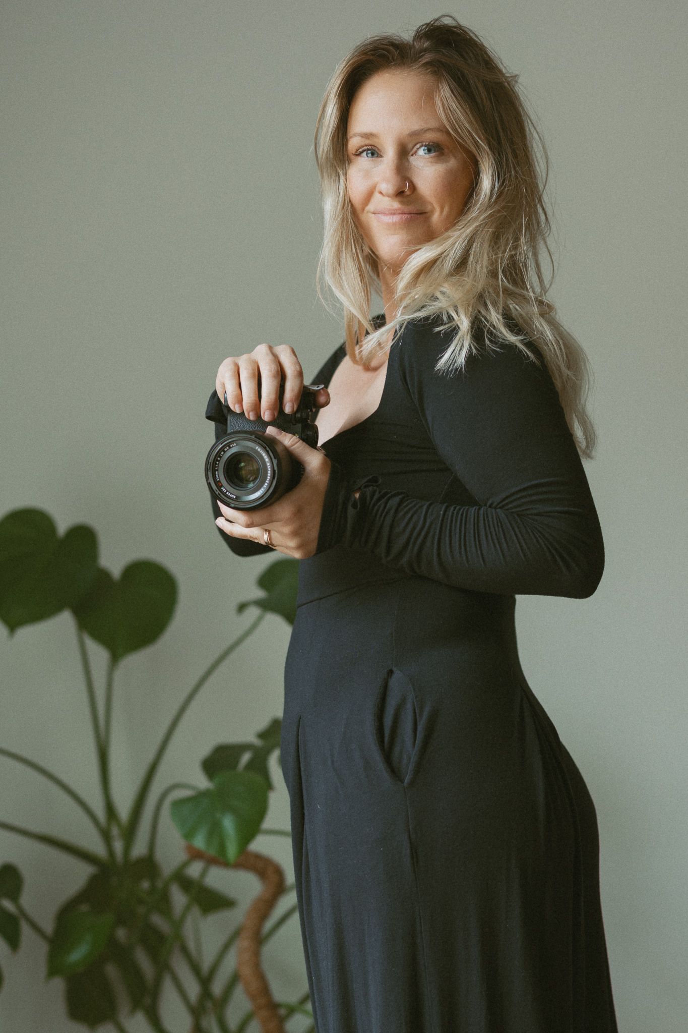 A smiling blonde woman in a black dress holds a camera, posing beside a green monstera plant.