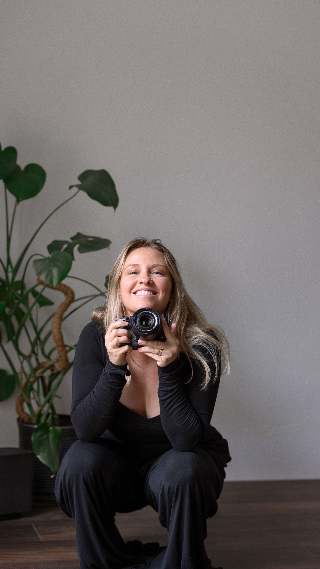 Smiling woman with long blonde hair, dressed in black, crouches while holding a camera beside a large monstera plant against a gray wall.