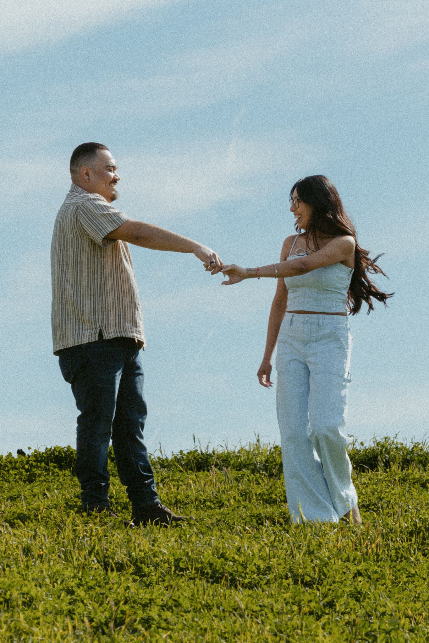 A couple holds hands and smiles at each other on a grassy hill under a blue sky at central coast winery