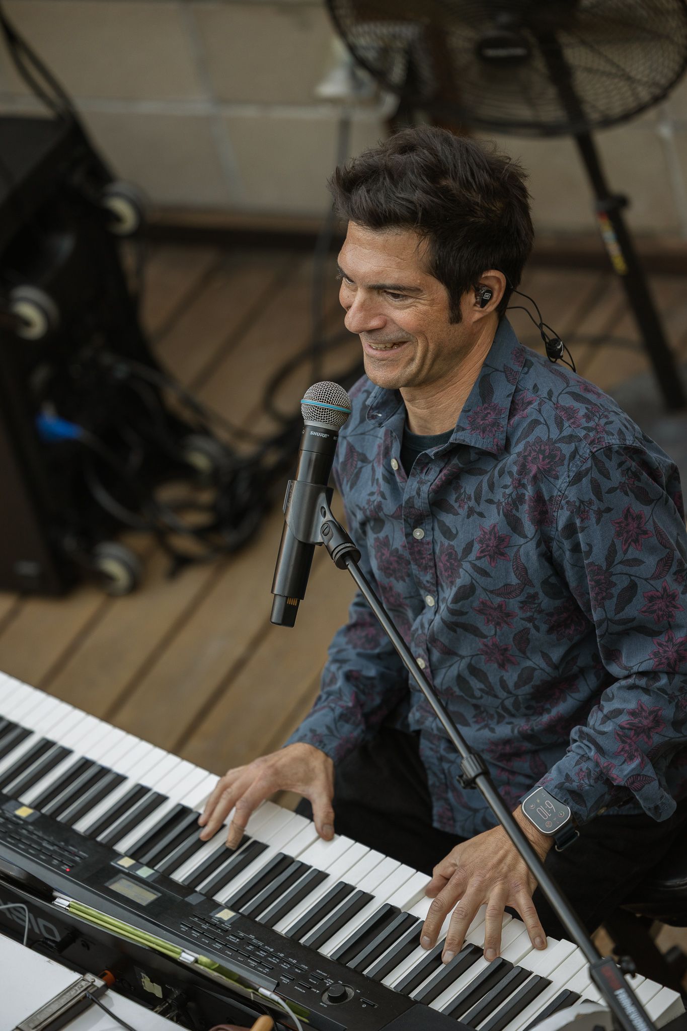 Smiling male musician playing keyboard synthesizer on stage, wearing floral shirt with microphone stand nearby.