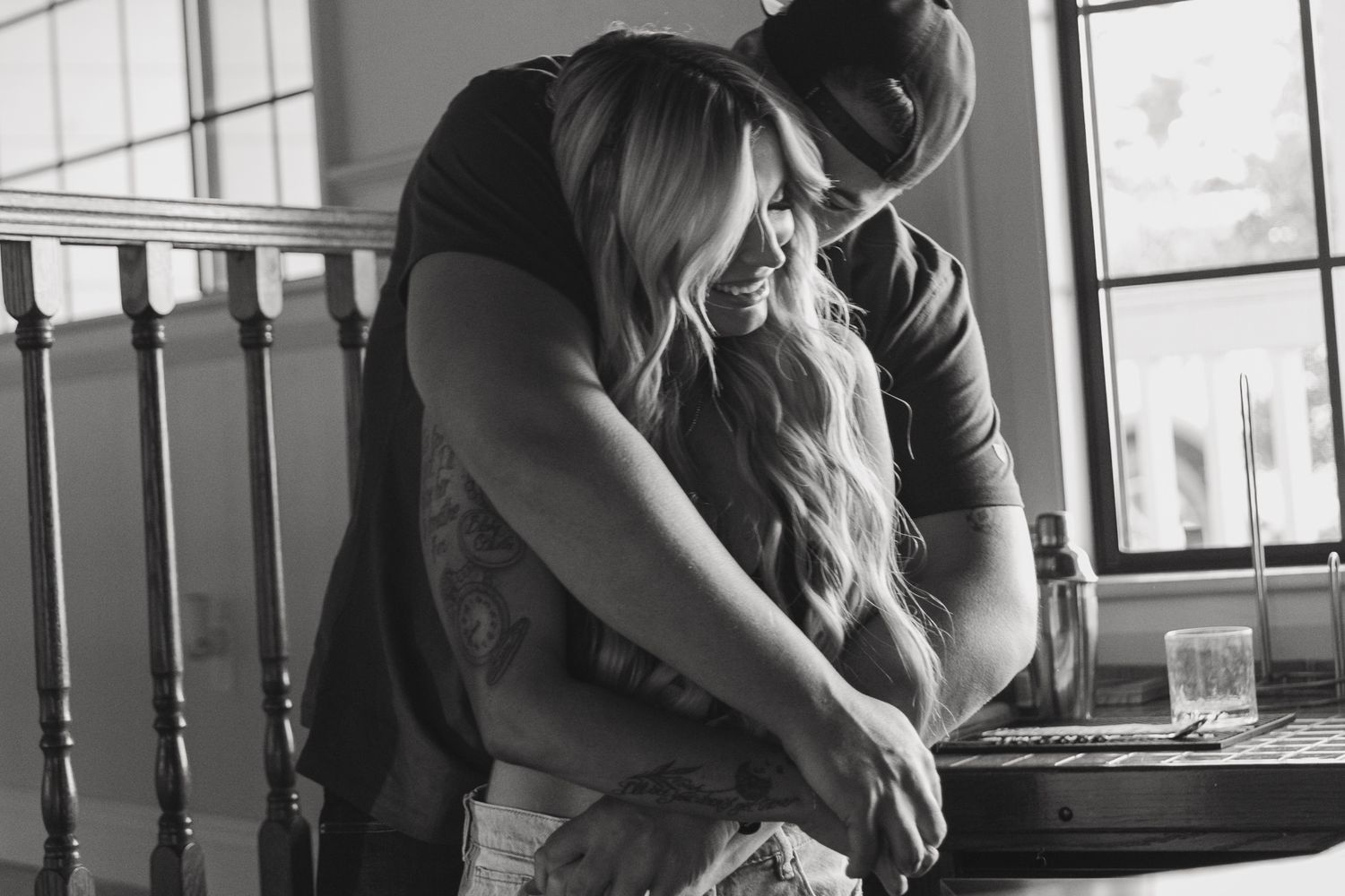 Black and white photo of a couple sharing an intimate embrace near a window indoors.