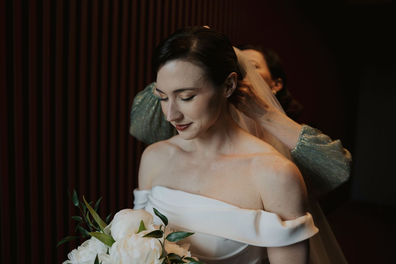 A bride in a white off-shoulder wedding gown adjusts her hair while getting ready for the ceremony.