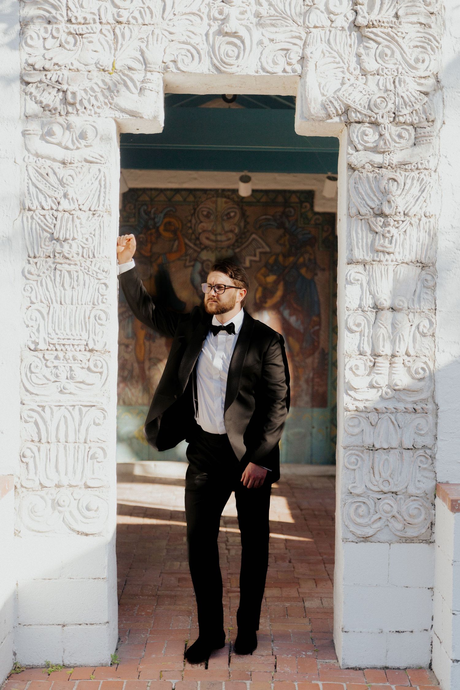 Dapper groom in black tuxedo and bow tie poses under ornate white carved archway.