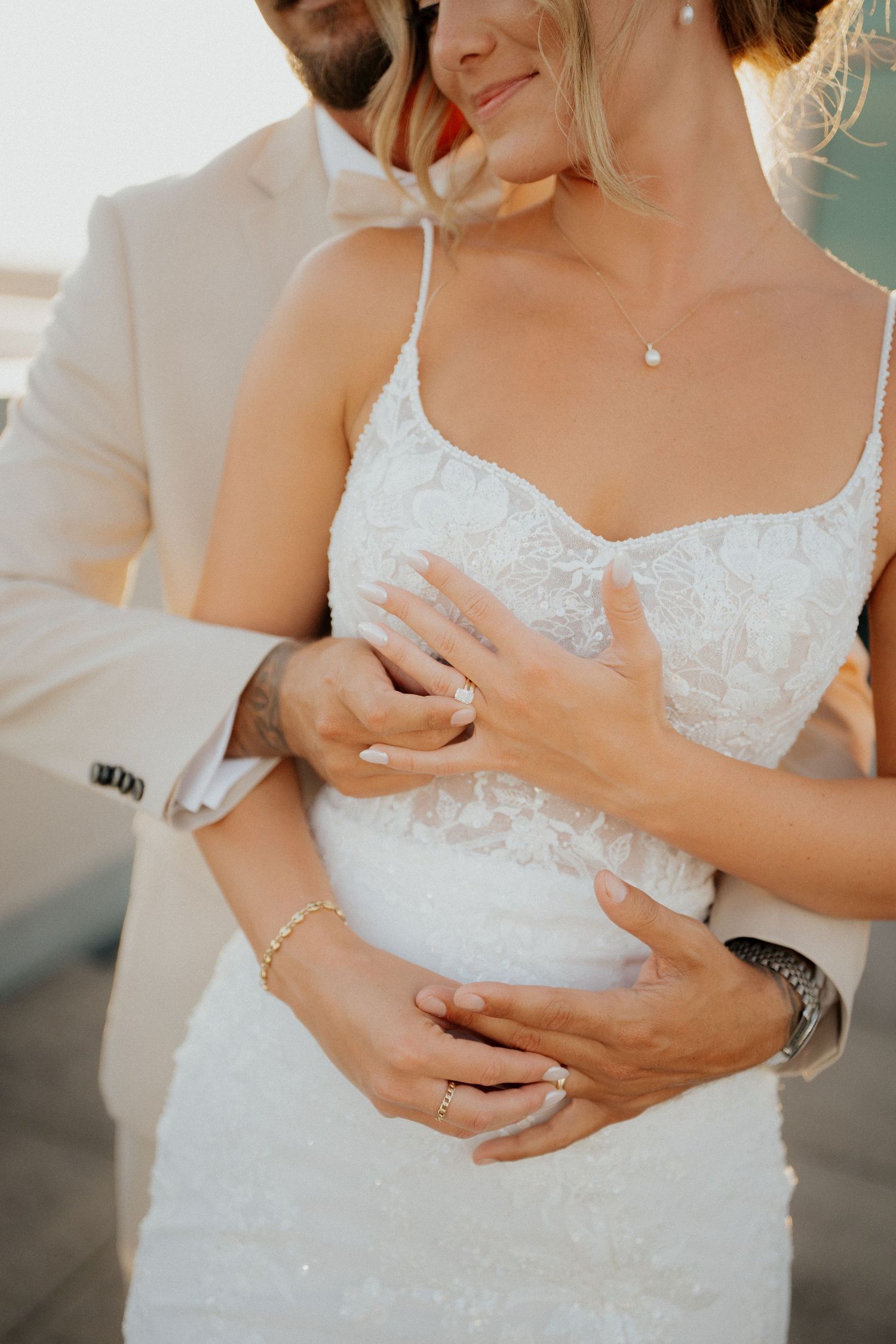 Close-up of bride in lace gown and groom in beige suit embracing, showcasing wedding rings and jewelry.