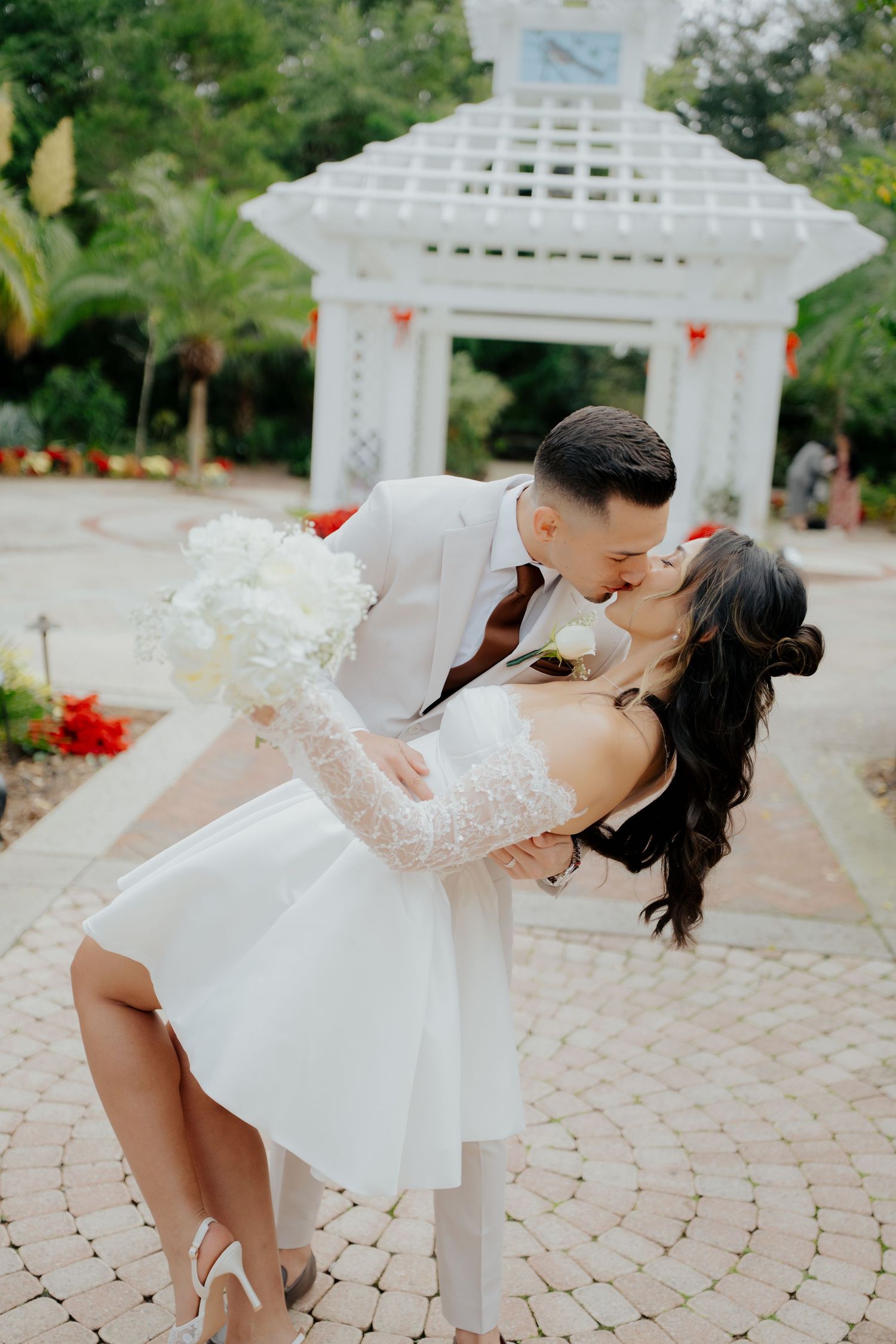Bride and groom share a romantic dip kiss in front of a white garden gazebo, bride holding white floral bouquet.