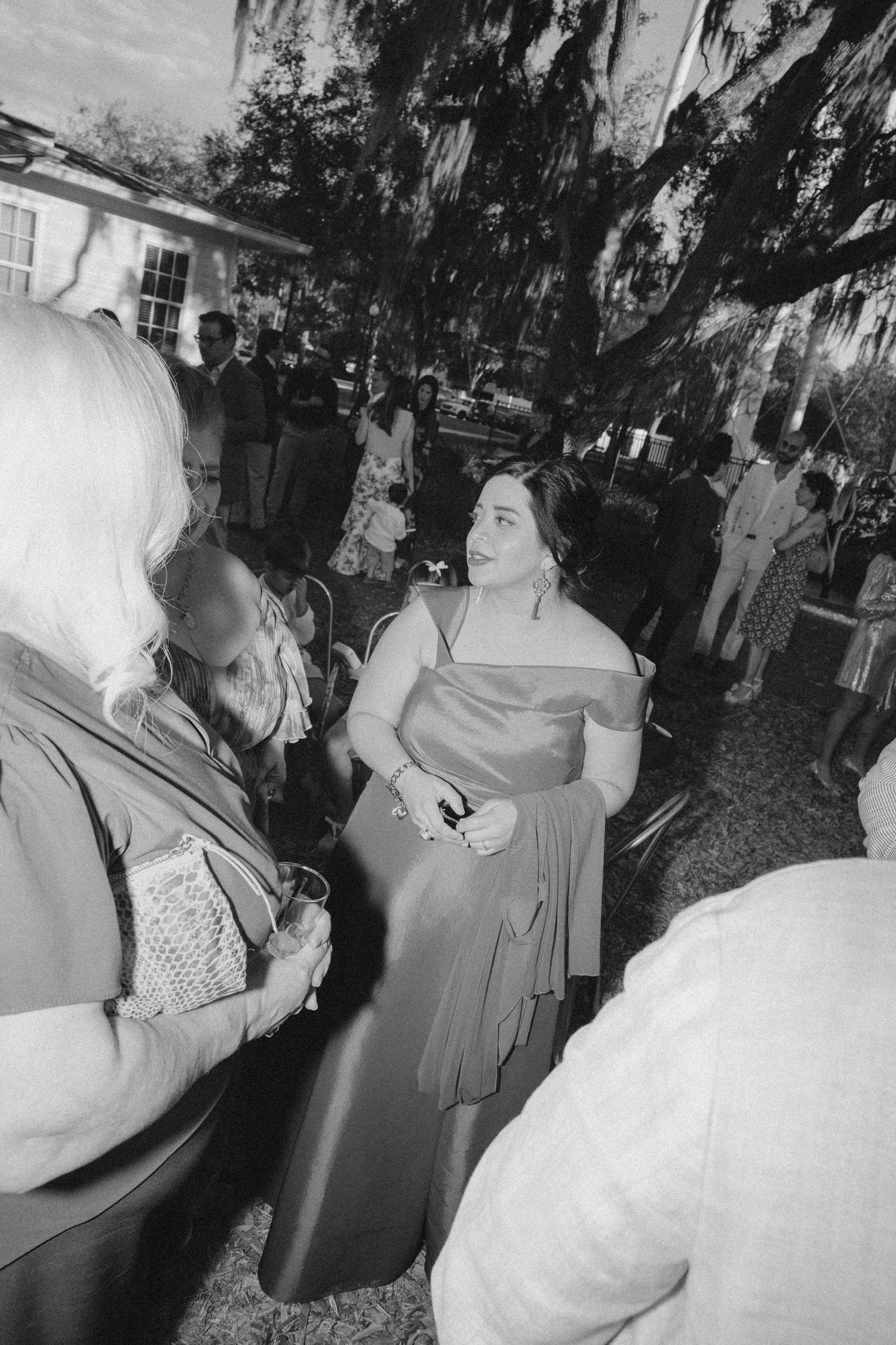 Black and white photo of woman in off-shoulder satin gown at outdoor wedding gathering.