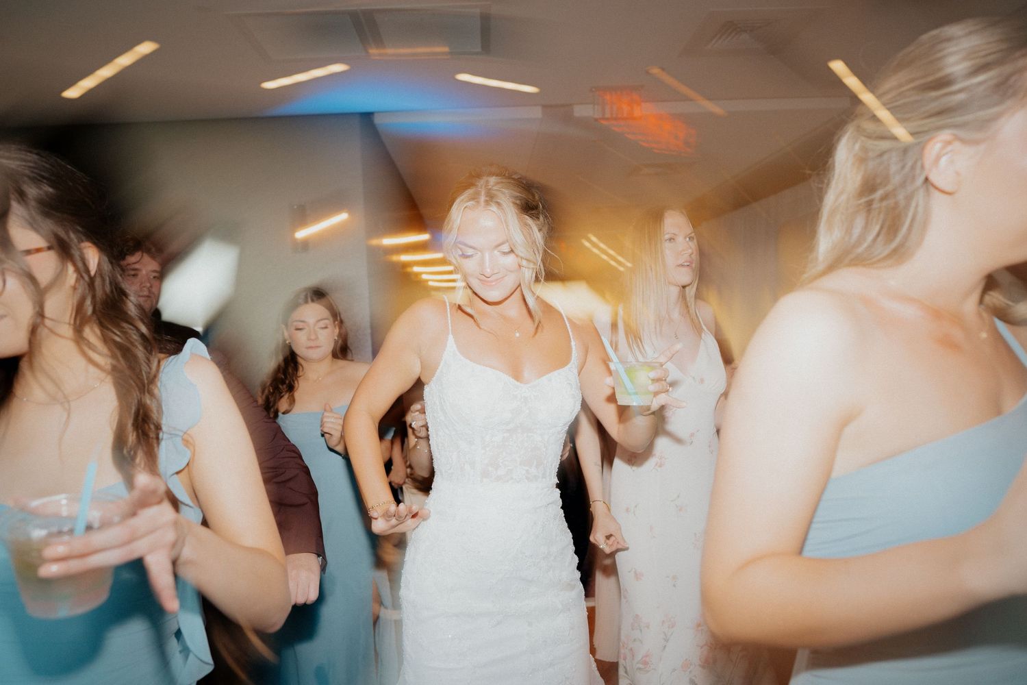 Bride in white lace gown dancing joyfully with guests at wedding reception under colorful lights.