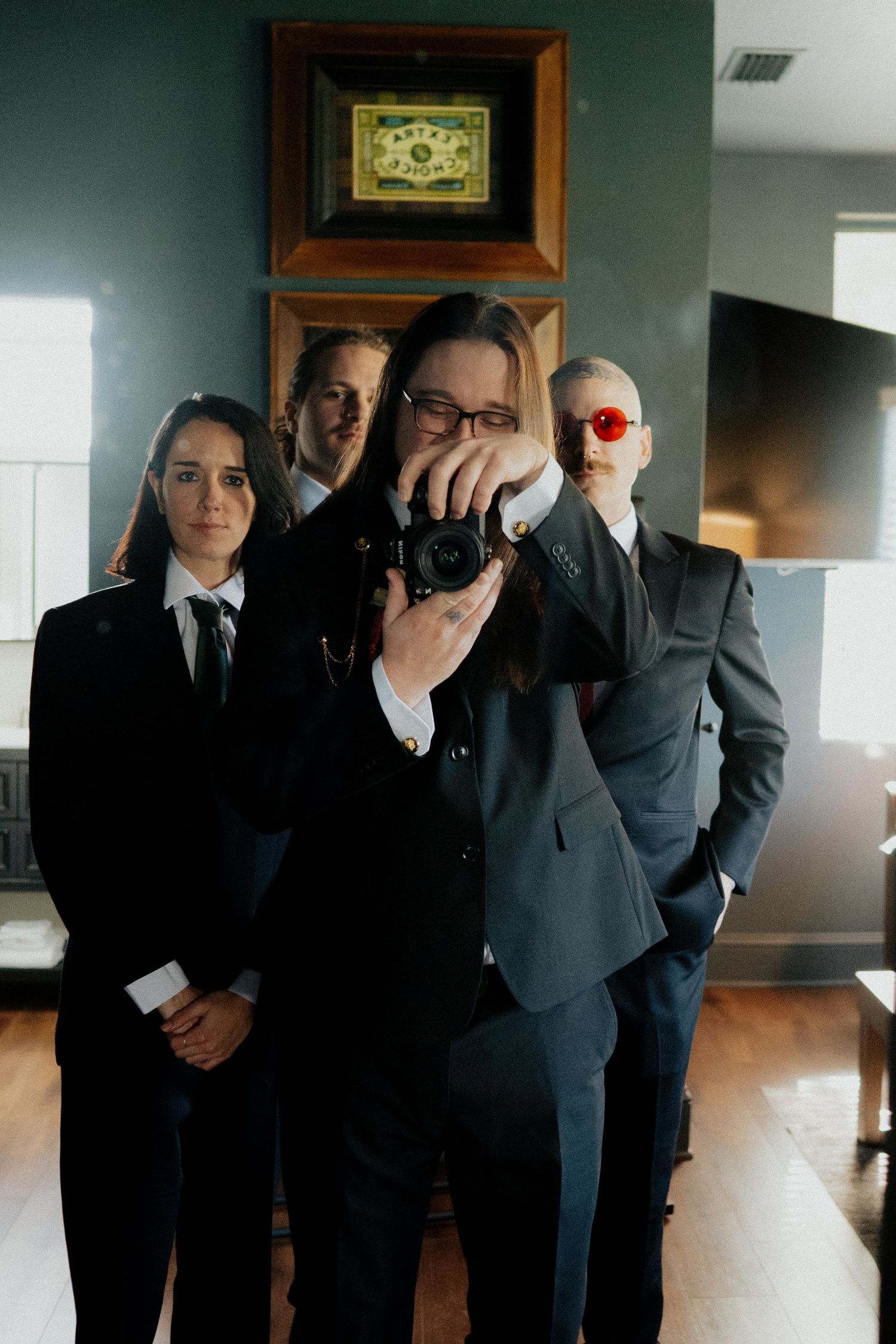 A group in formal black suits stand in a row indoors while covering their eyes with their hands.