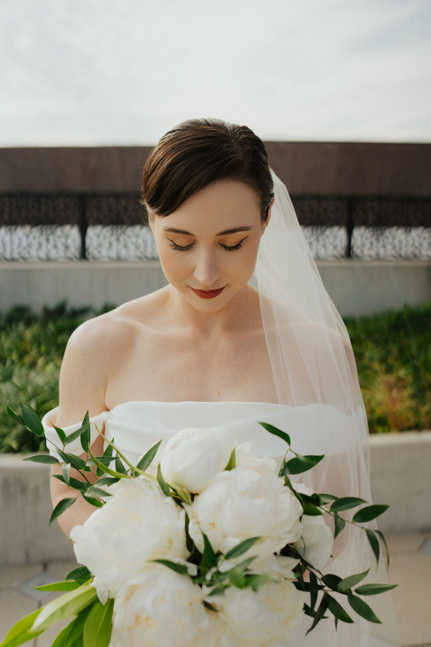 Close-up of a bride holding an elegant white peony bouquet with green foliage against a light background.