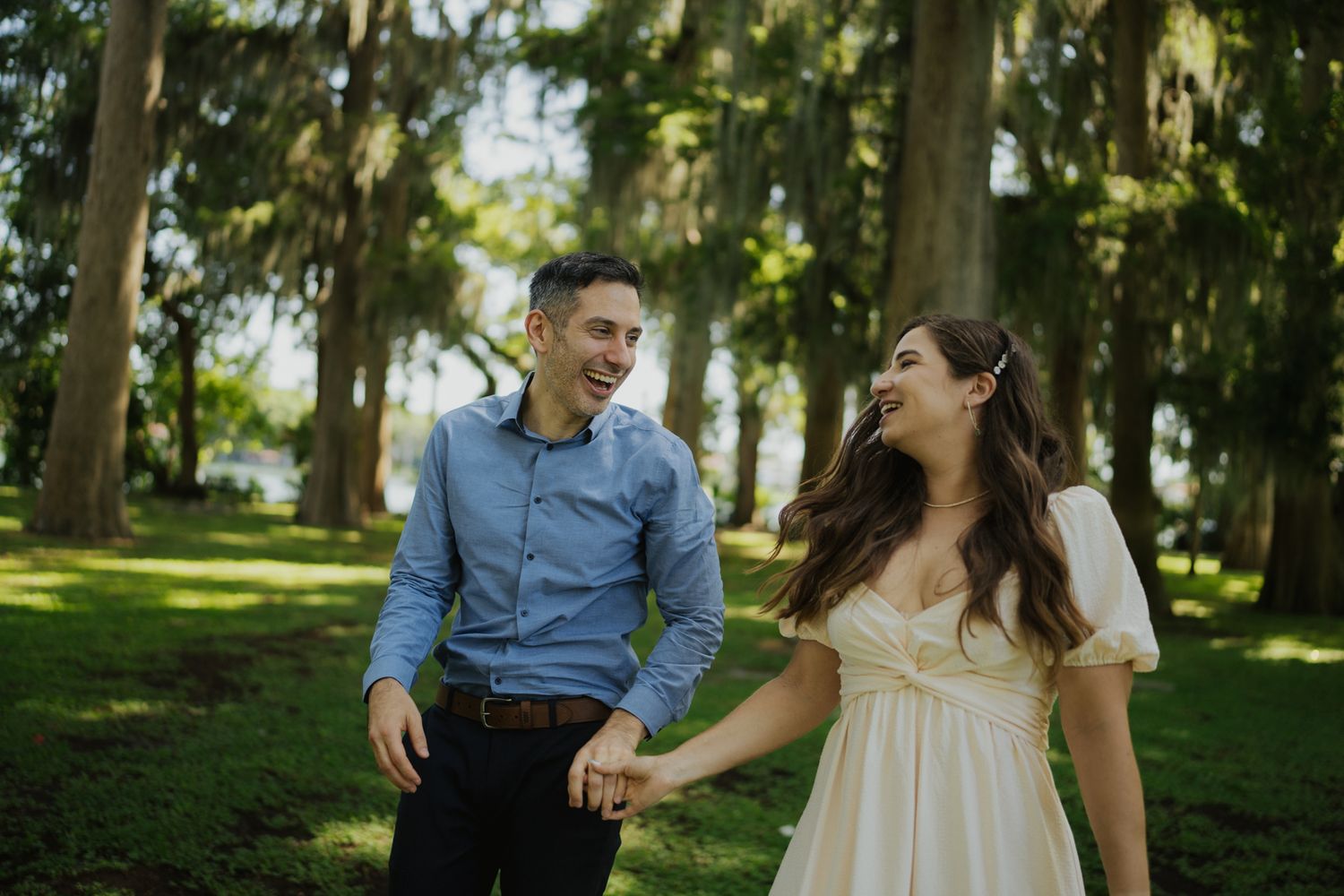 A couple laughs together while holding hands in a sunlit park with tall trees.
