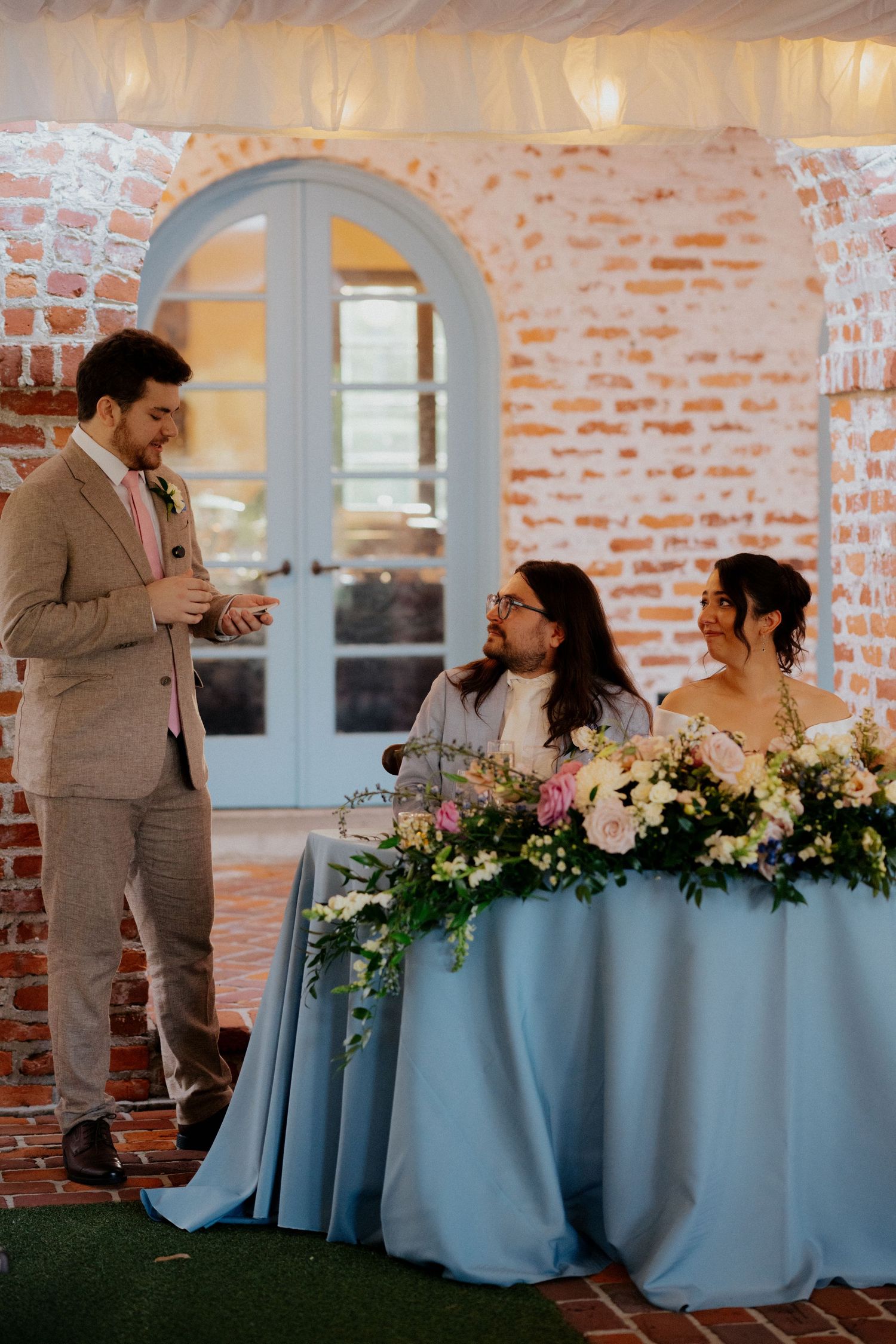 Wedding reception at a rustic brick venue with light blue tablecloth and floral arrangements during a speech.