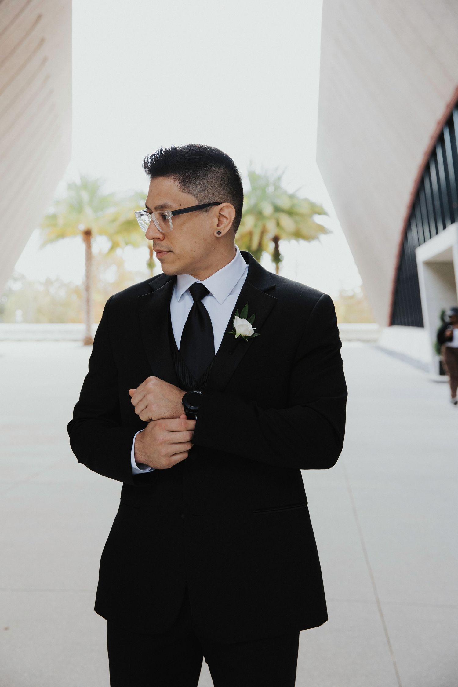 Groom in classic black suit and tie adjusts cuff while standing near white columns with palm trees in background.