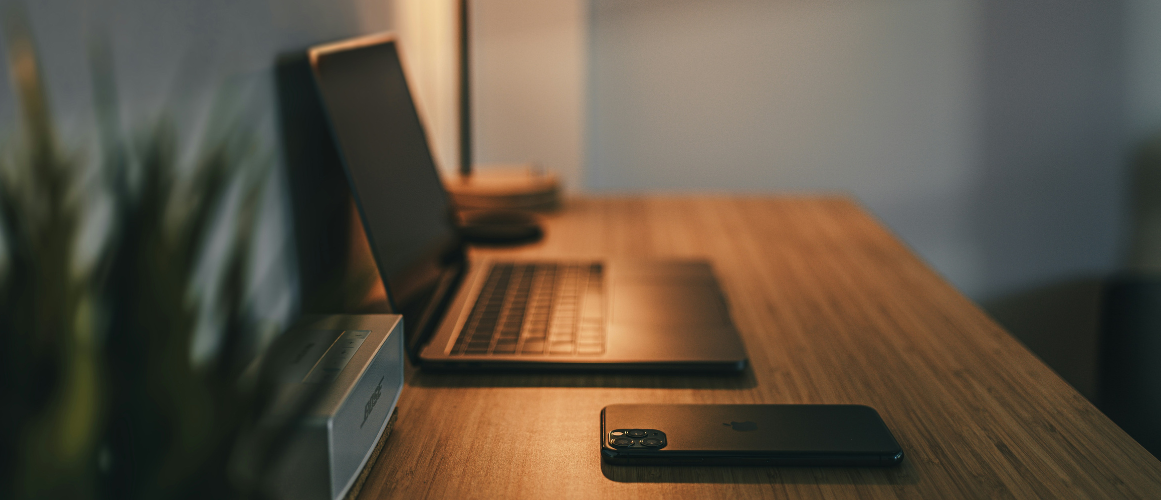 A minimalist workspace with laptop and phone on wooden desk in warm lighting.
