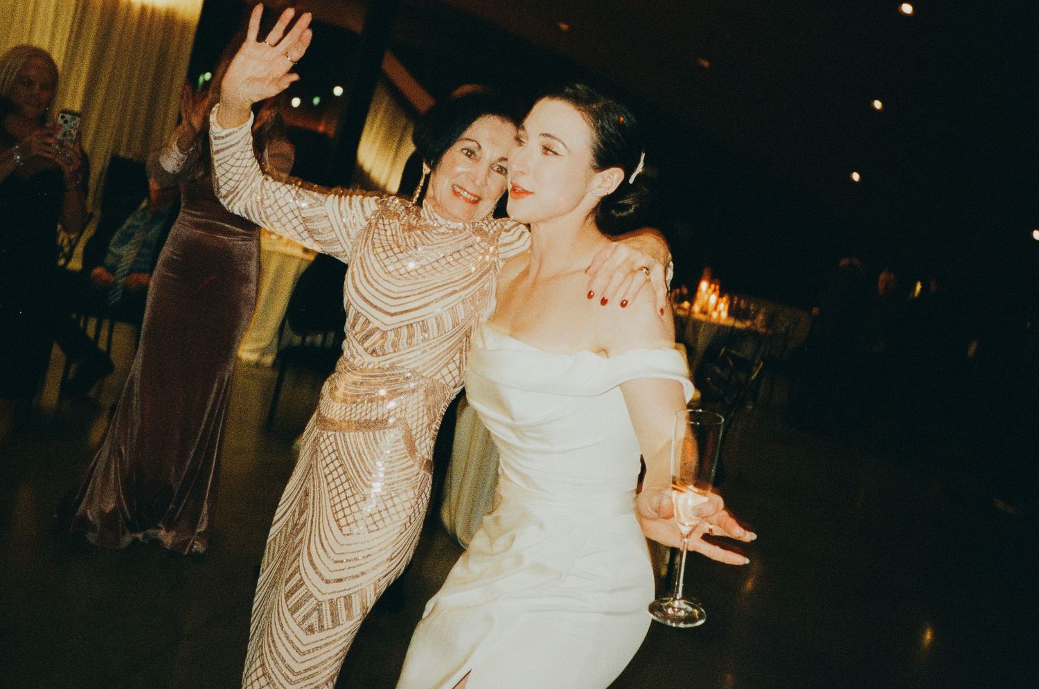 Joyful bride in white off-shoulder gown dancing with a guest in a beaded dress at wedding reception.