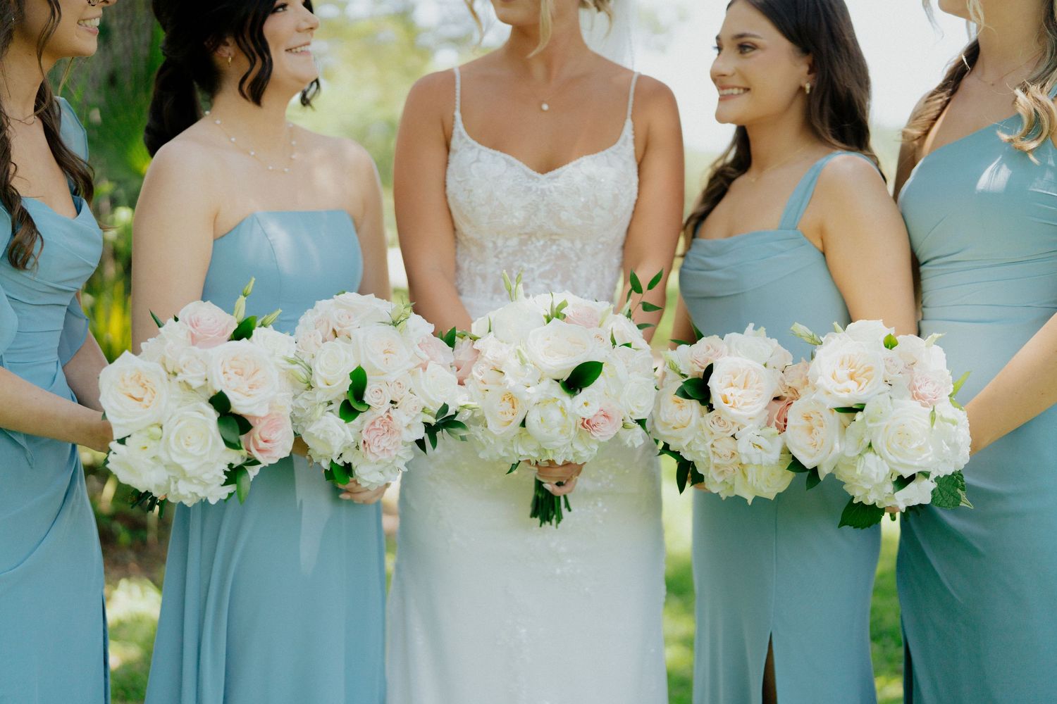 Bride in lace gown with bridesmaids in blue dresses holding white and pink bouquets outdoors.