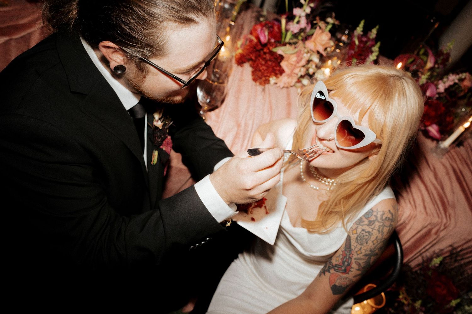 Groom feeds bride wedding cake as she wears heart-shaped white sunglasses at reception.