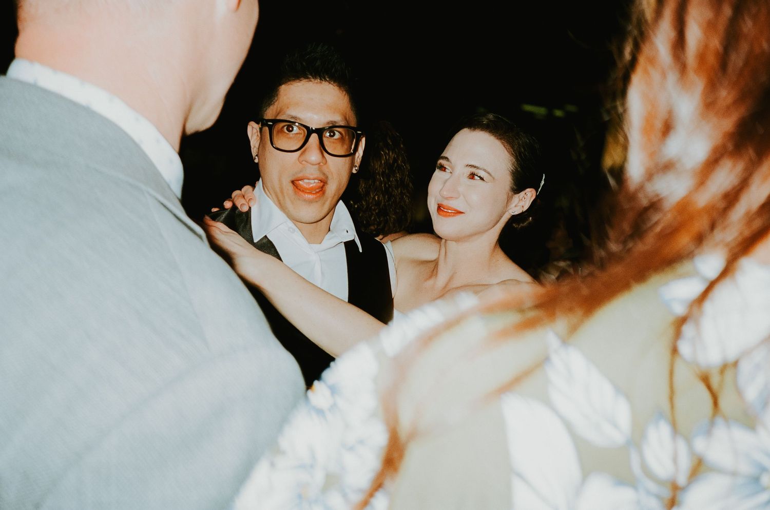Couple at a wedding reception, man in glasses and woman in white dress socializing.