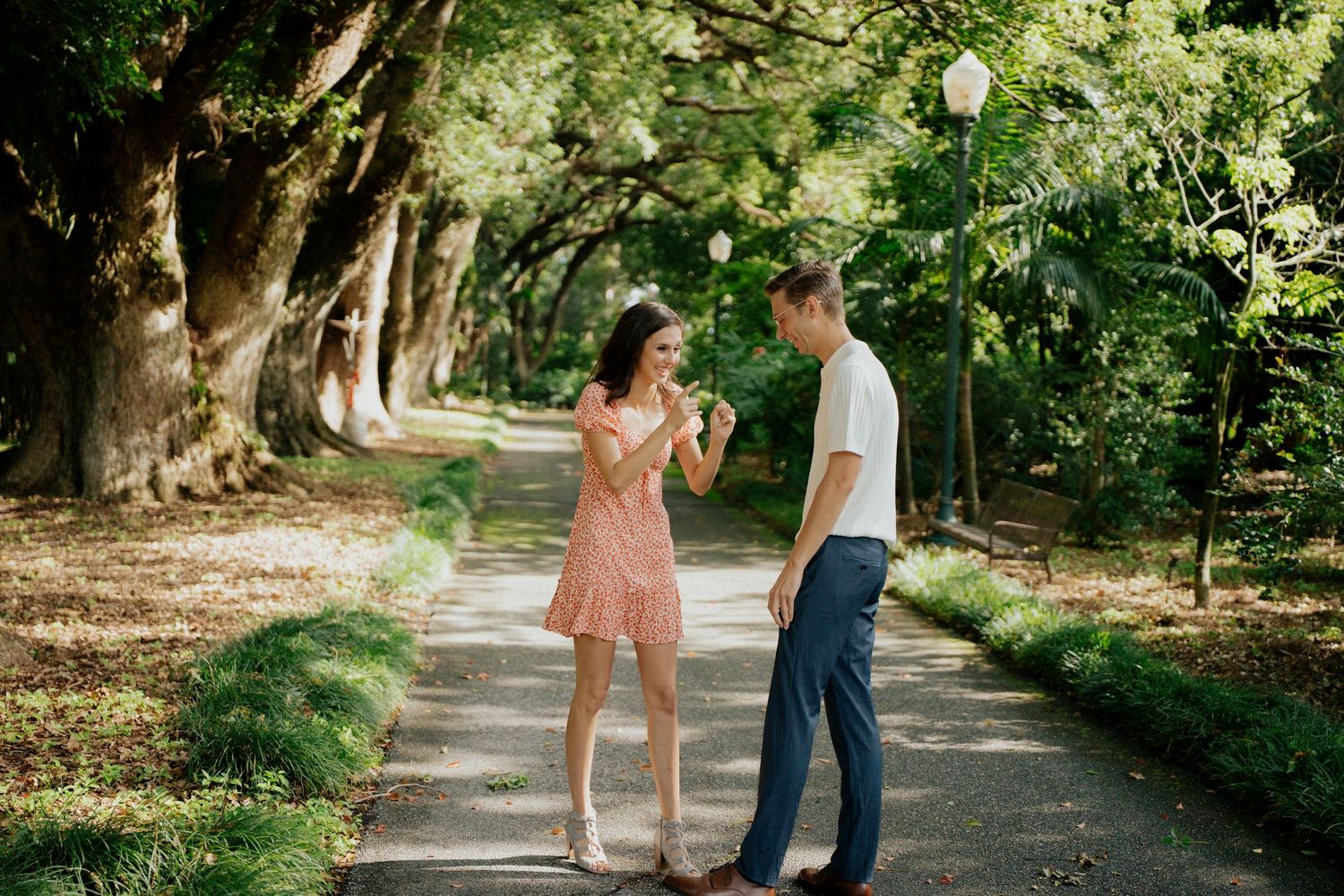 Couple in romantic engagement photo session walking along a tree-lined path in a lush green park setting.