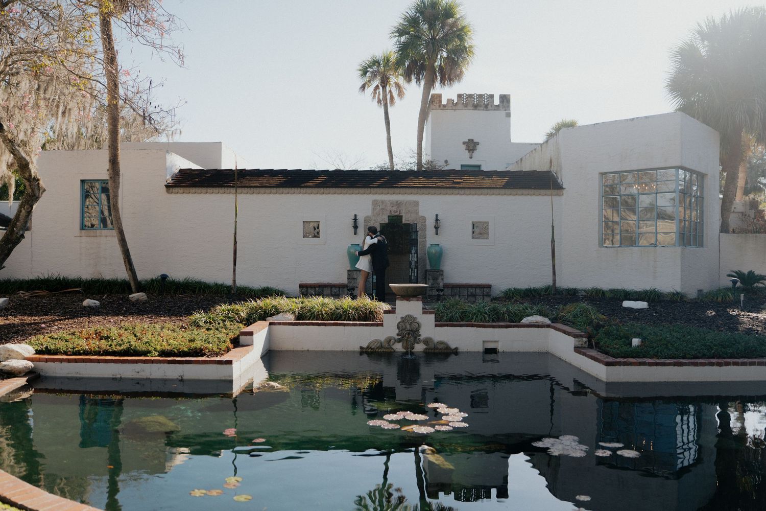 Spanish-style white building with reflecting pool and garden beds in a serene courtyard setting with palm trees.
