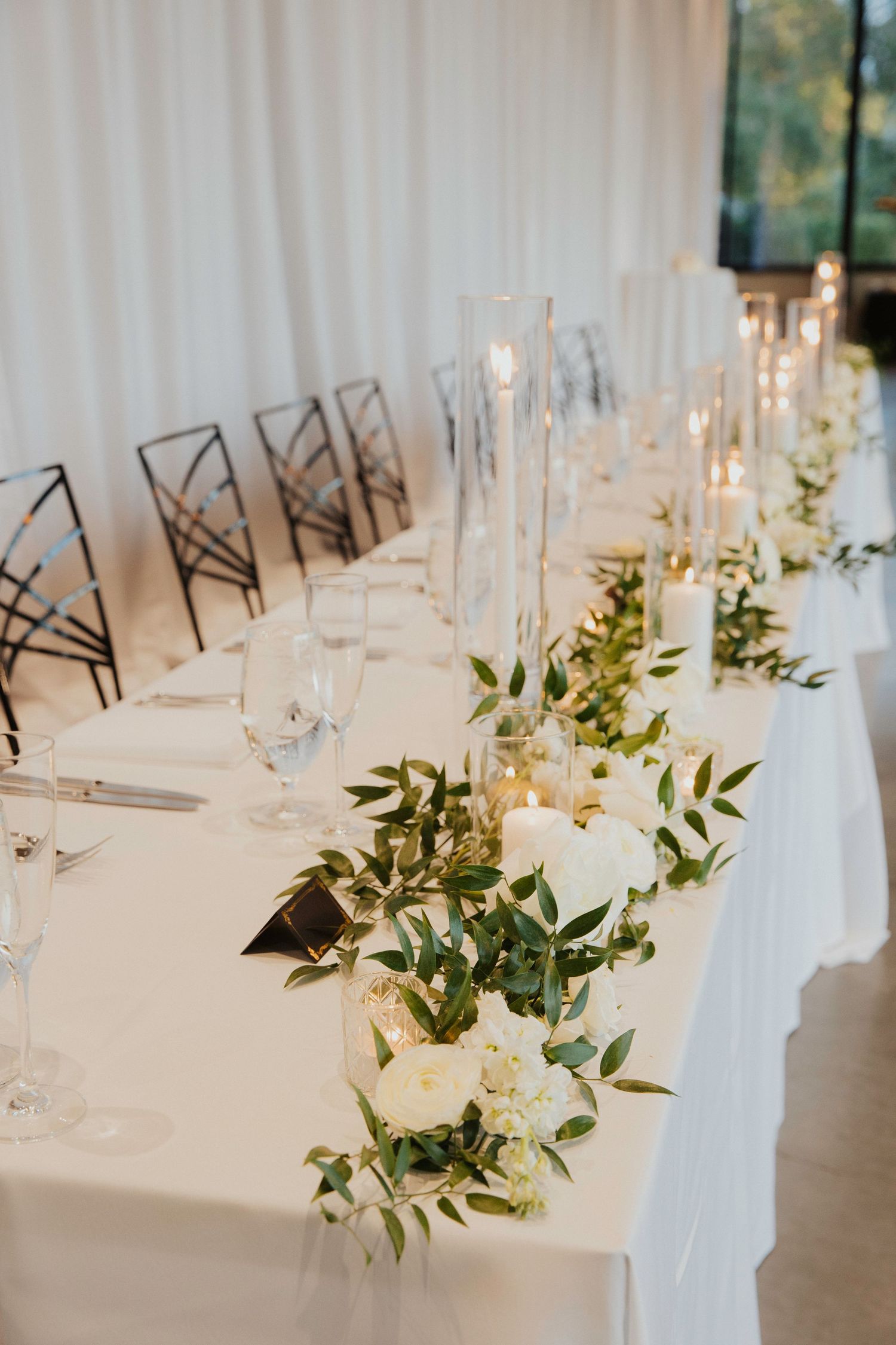 Elegant wedding reception table setting with white linens, glassware and floral centerpieces with greenery.