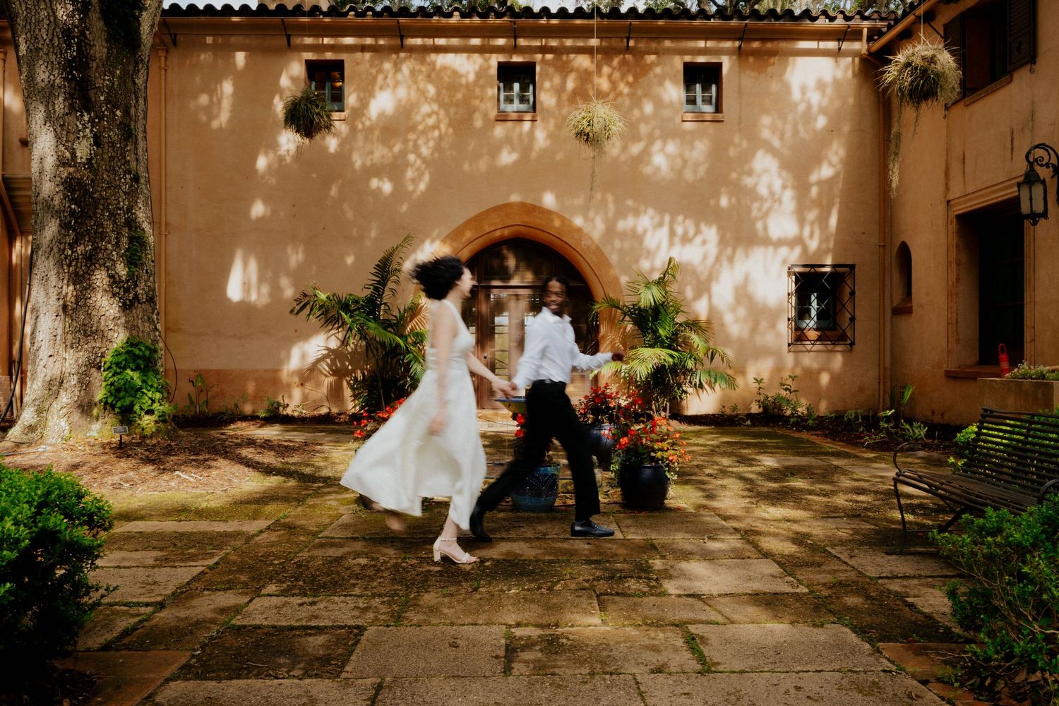 Joyful couple dancing in a sunlit courtyard of a historic Spanish-style building with floral decor.