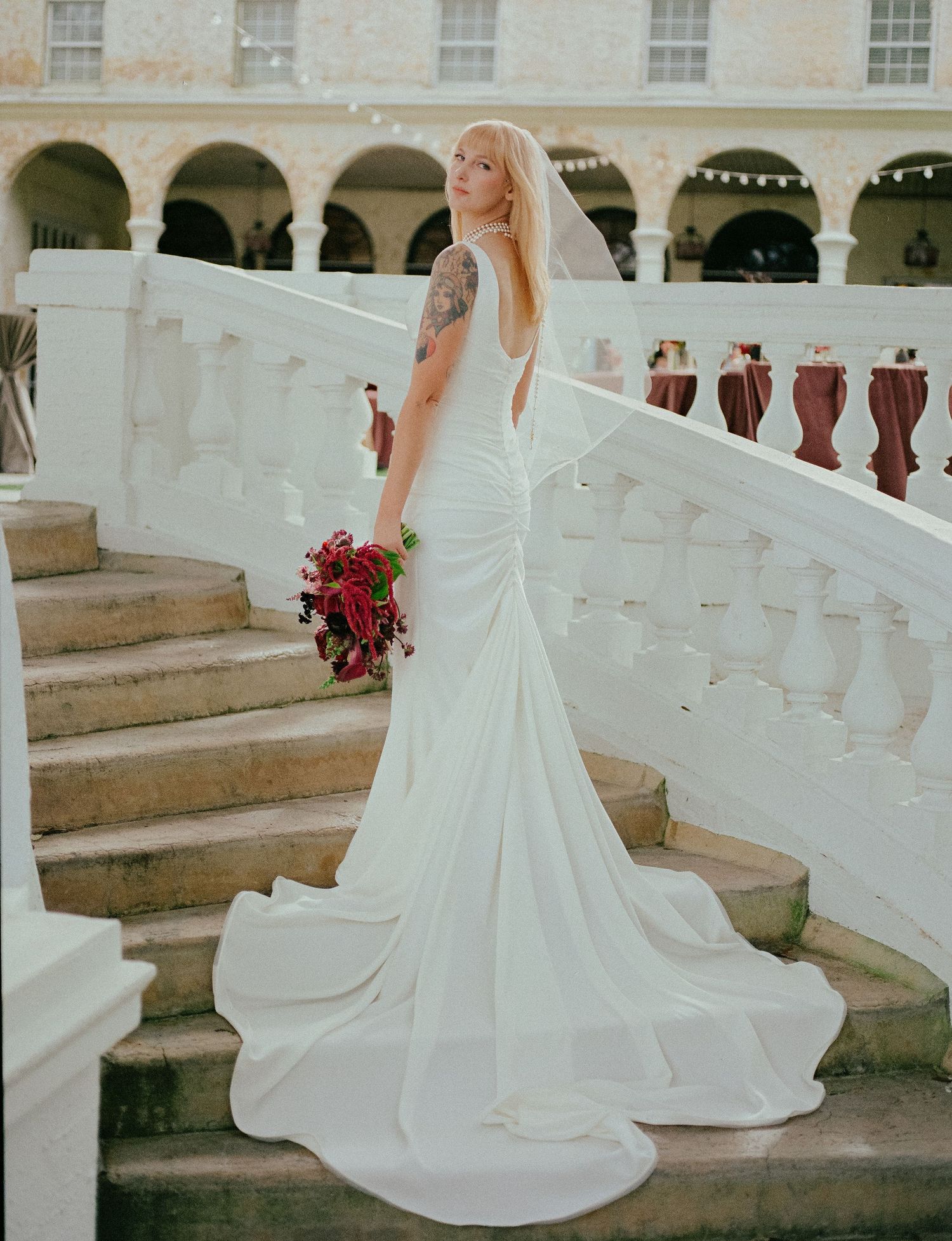 Blonde bride with tattoo in fitted white gown on grand staircase with red bouquet.